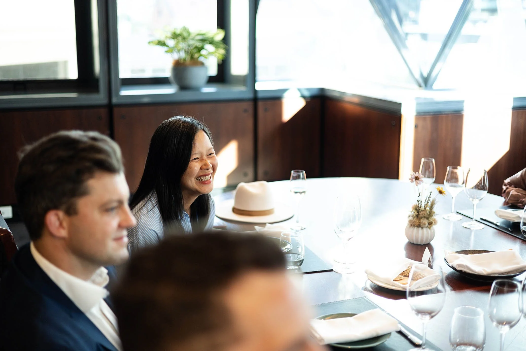 People sitting around a dining table, smiling and engaging in conversation, in a well-lit room with large windows, a potted plant, and a hat on the table.