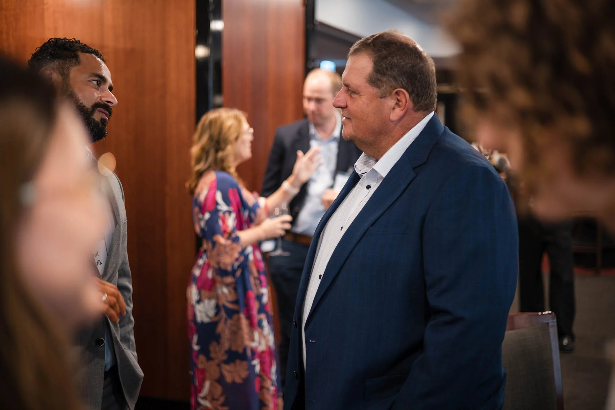 A man in a blue suit smiling and talking to another man at a social event. Other people are engaged in conversations in the background.