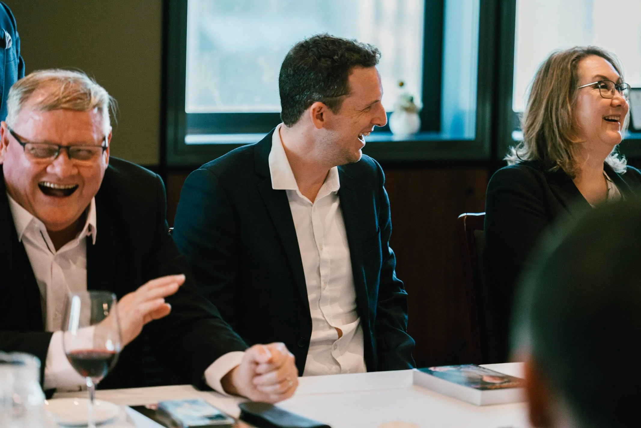 People sitting around a table, laughing and enjoying a moment during a meeting or gathering, with wine glasses and books on the table, in a well-lit room.
