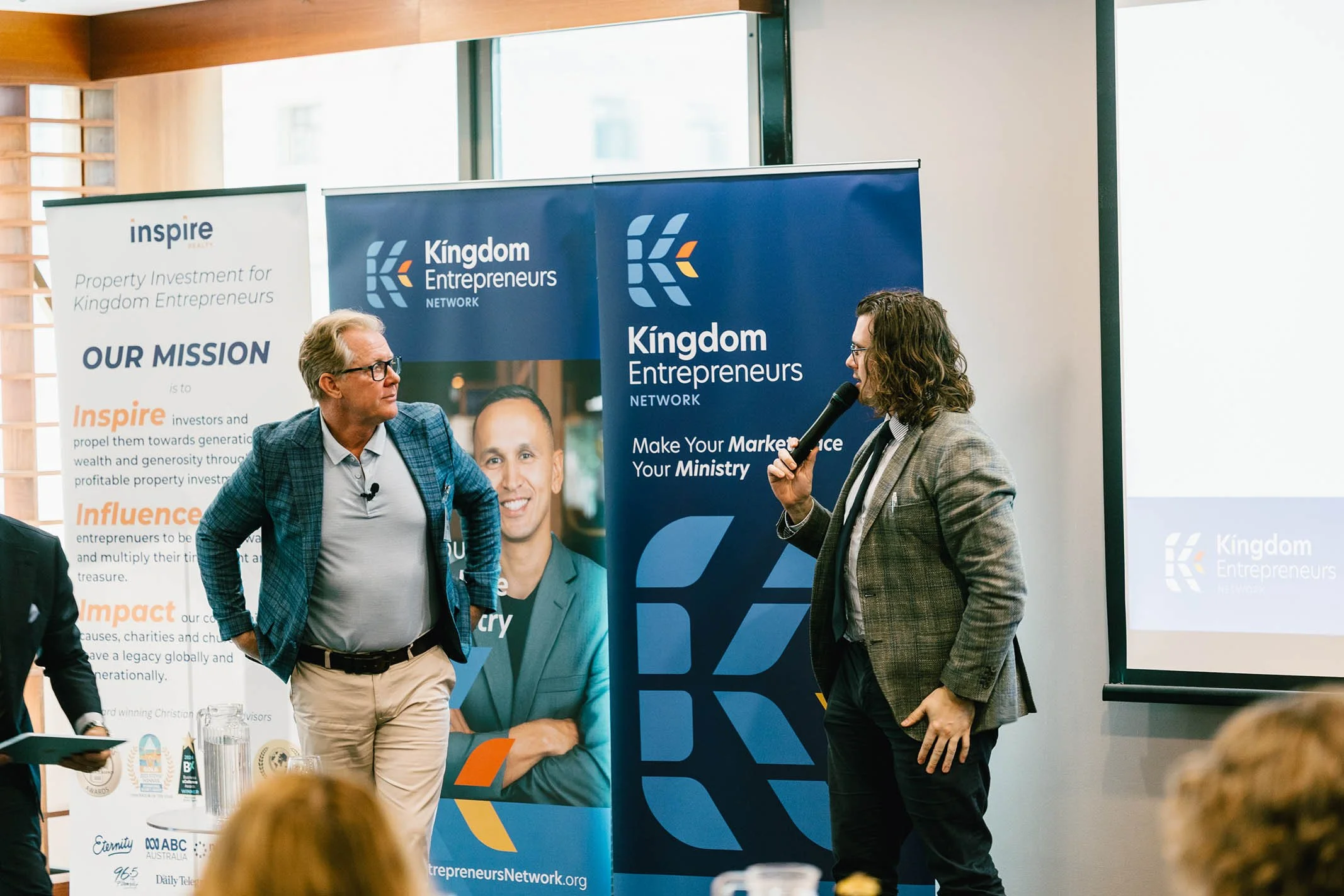Two men at a business event, one speaking into a microphone and the other listening, with promotional banners behind them for Kingdom Entrepreneurs Network and Inspire, in a conference room.