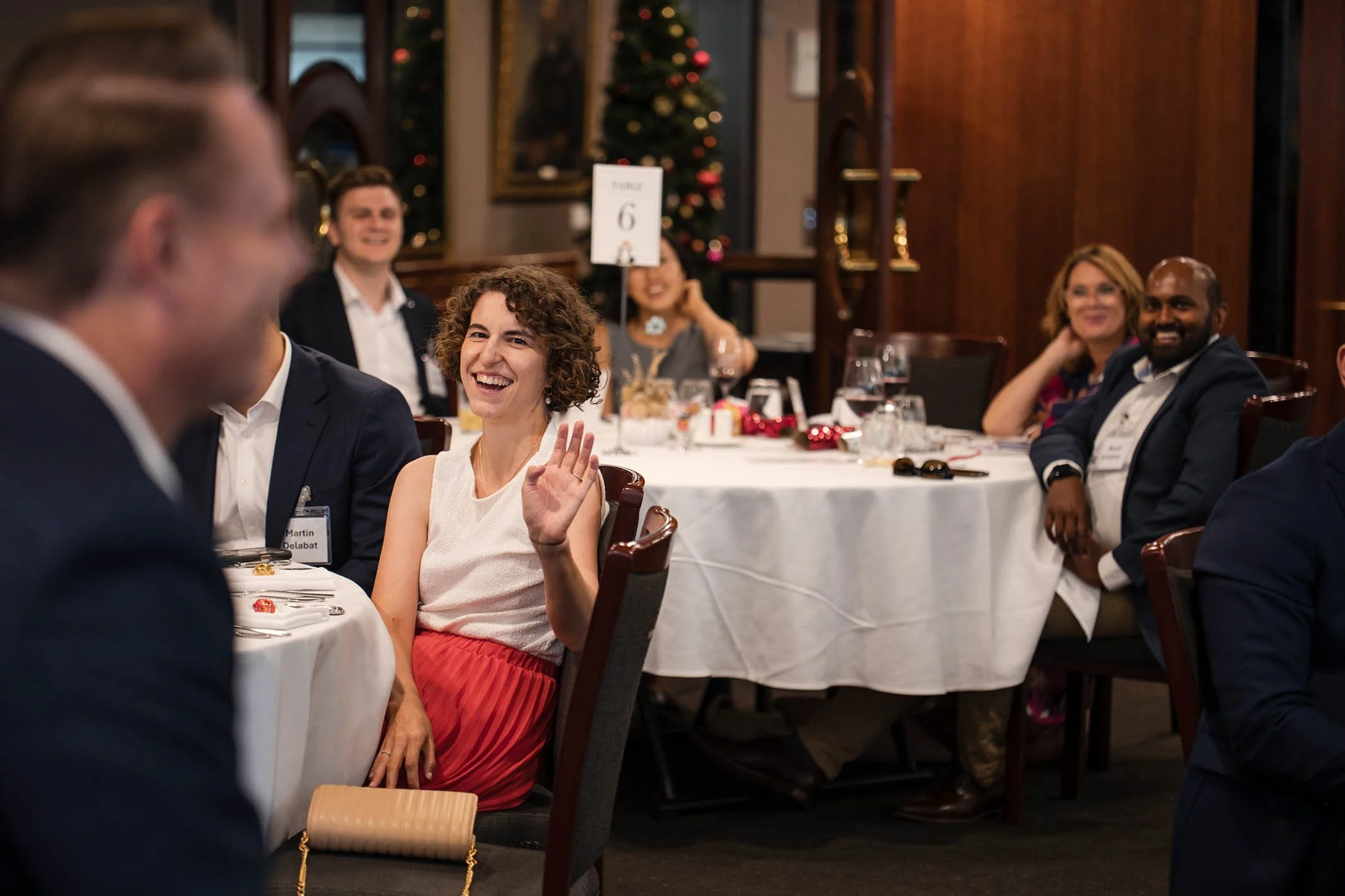 A woman at a formal dinner event waving and smiling, with several other people in the background seated around tables decorated for the holidays, including a Christmas tree.