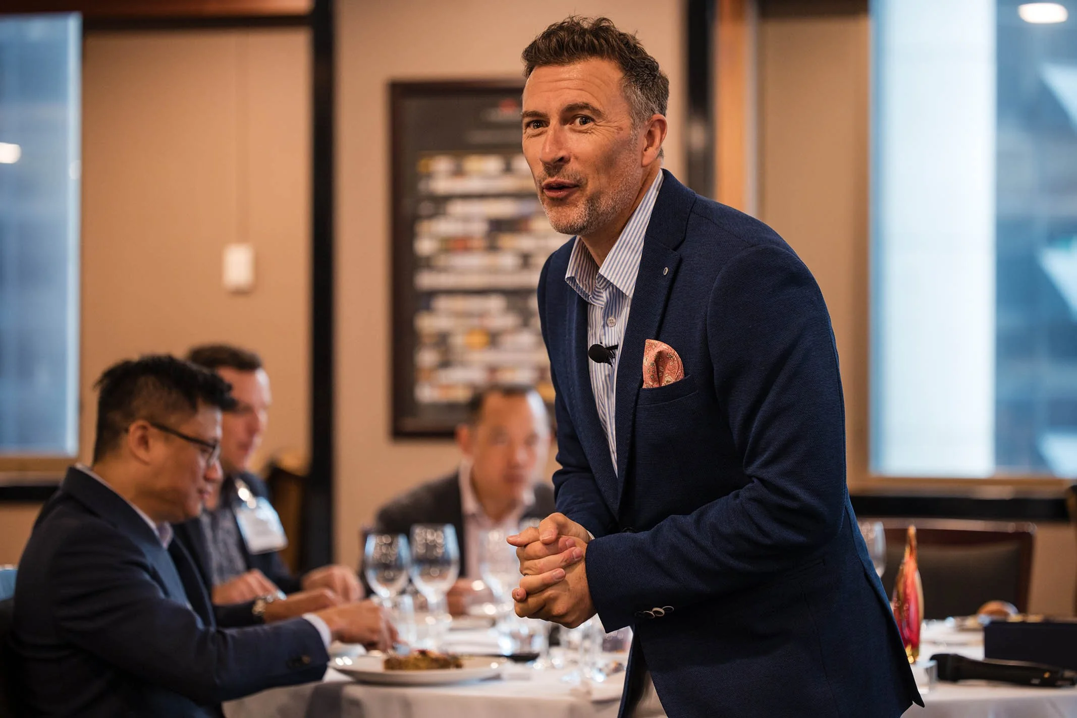 A man in a blue suit speaking at a conference, with seated attendees in the background.