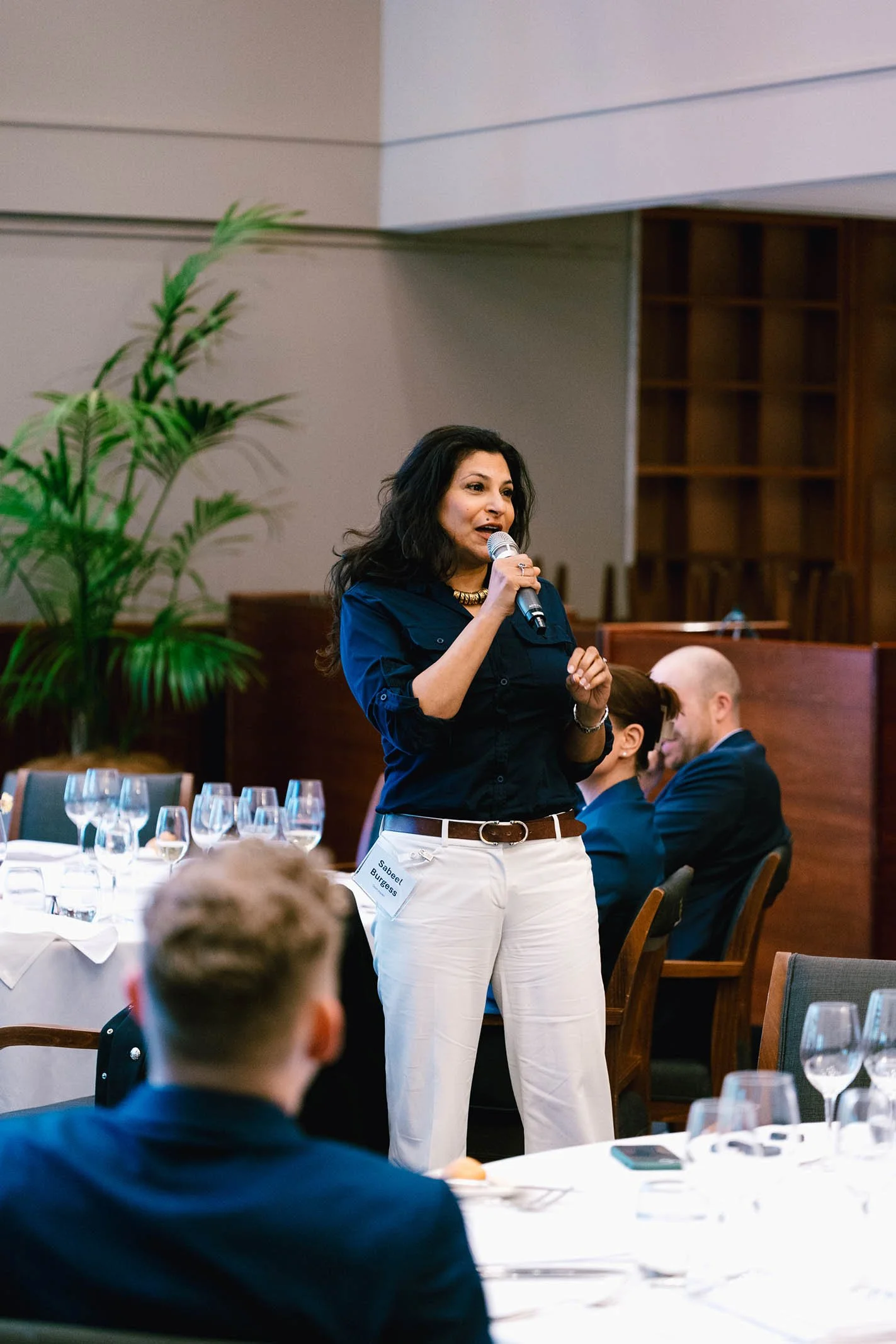 A woman speaking into a microphone at a formal event, with seated attendees and wine glasses on a table in the background.