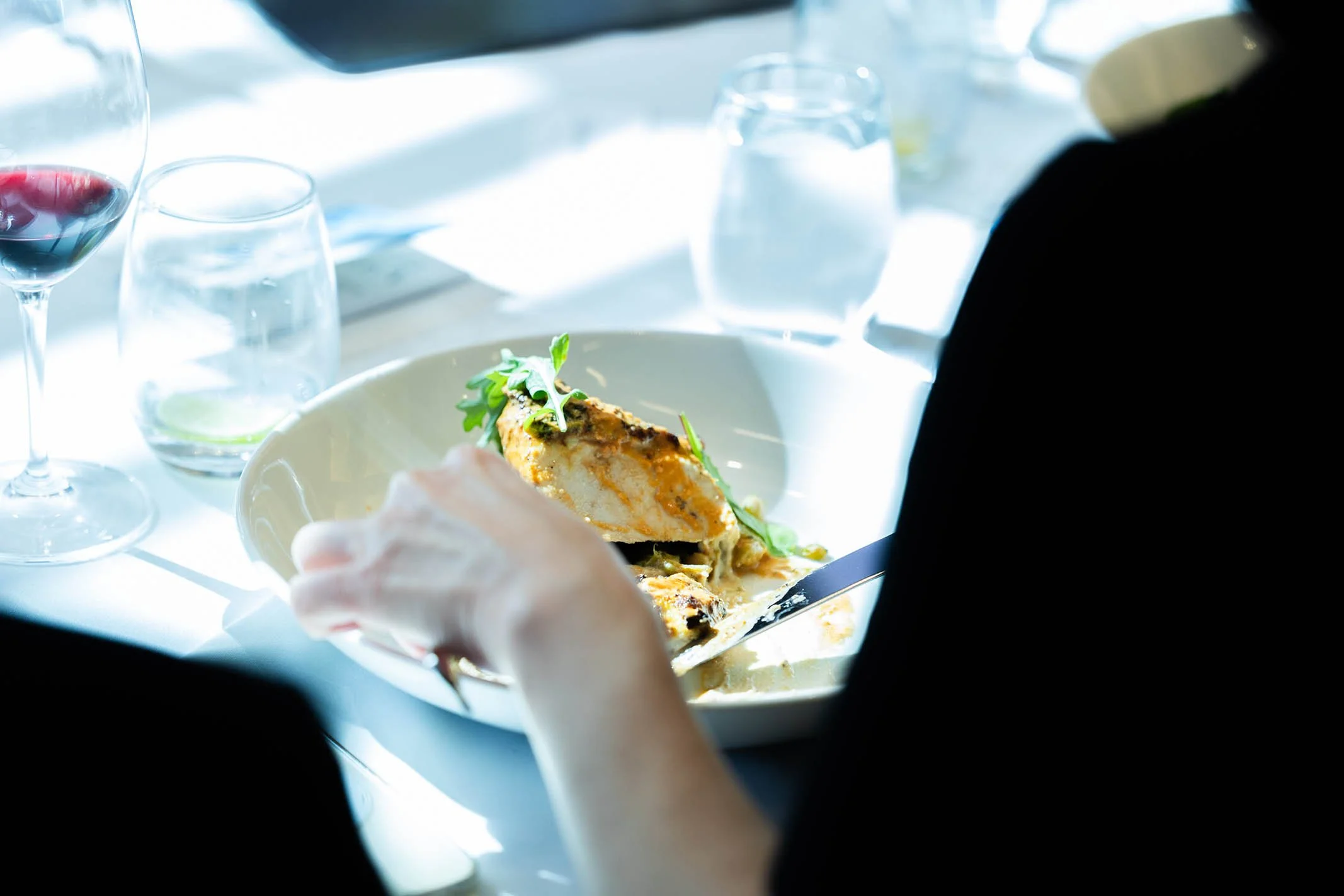 Person cutting food on a white plate at a dining table with glasses of water and wine.