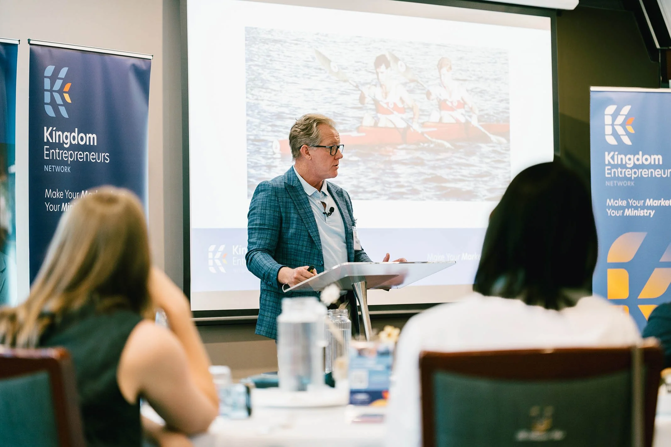 A man giving a presentation in front of a screen and banners with the logo and name 'Kingdom Entrepreneurs Network' at a conference.