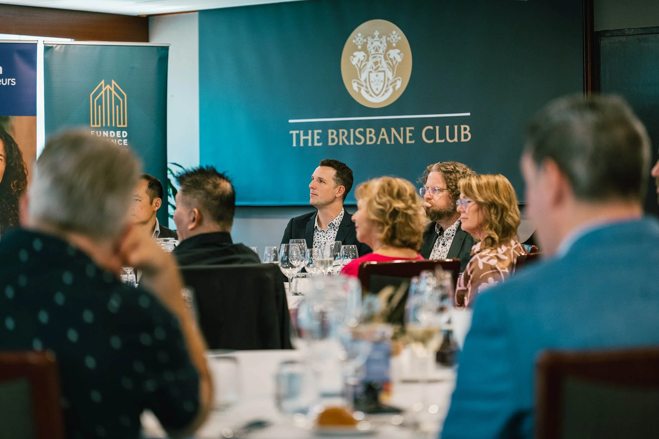 People attending a formal event at The Brisbane Club, seated at tables with wine glasses, listening to a speaker or presentation, with a large sign and crest on the wall behind them.