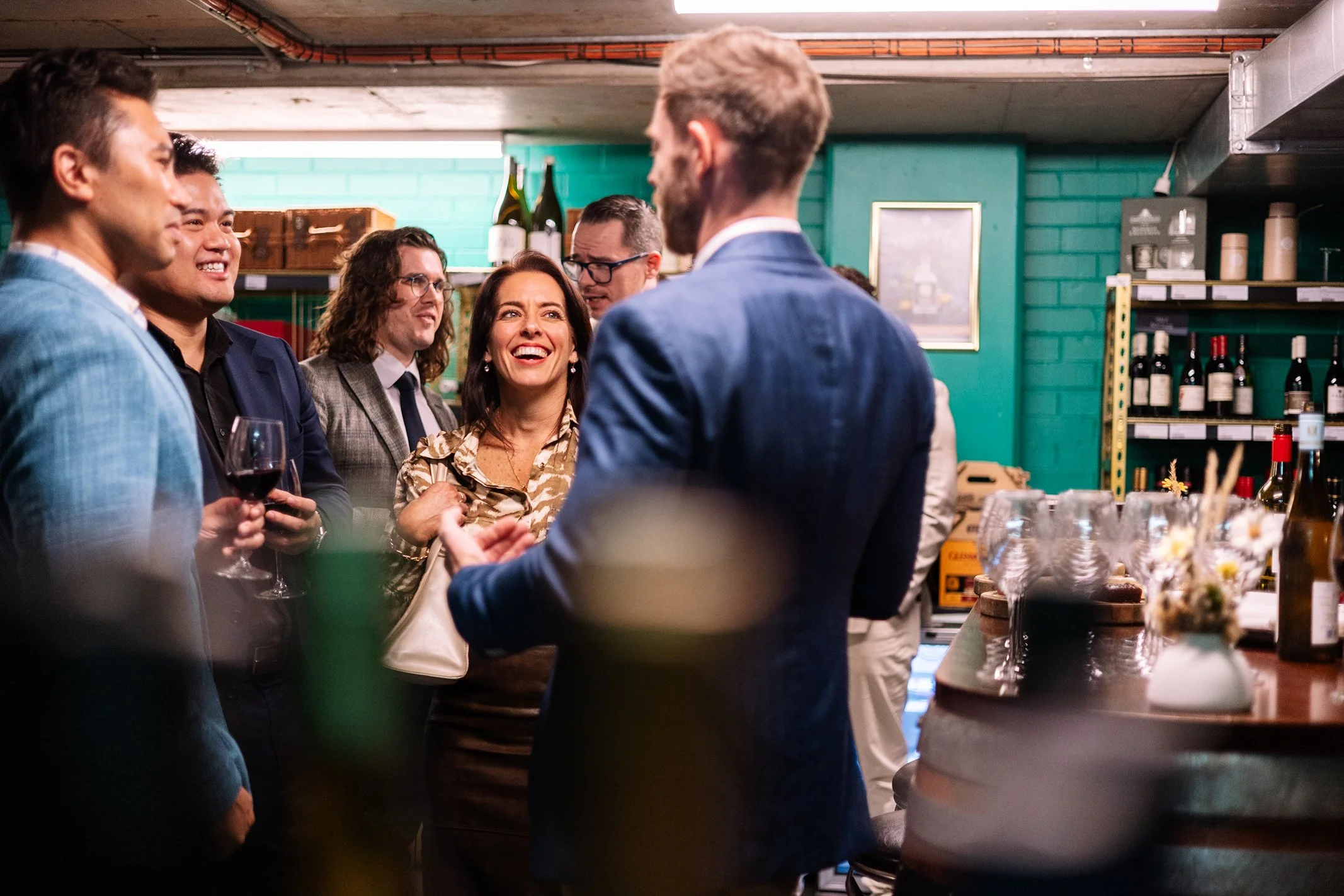 Group of people at a social gathering, talking and smiling indoors with wine bottles and glasses on shelves and tables in the background.