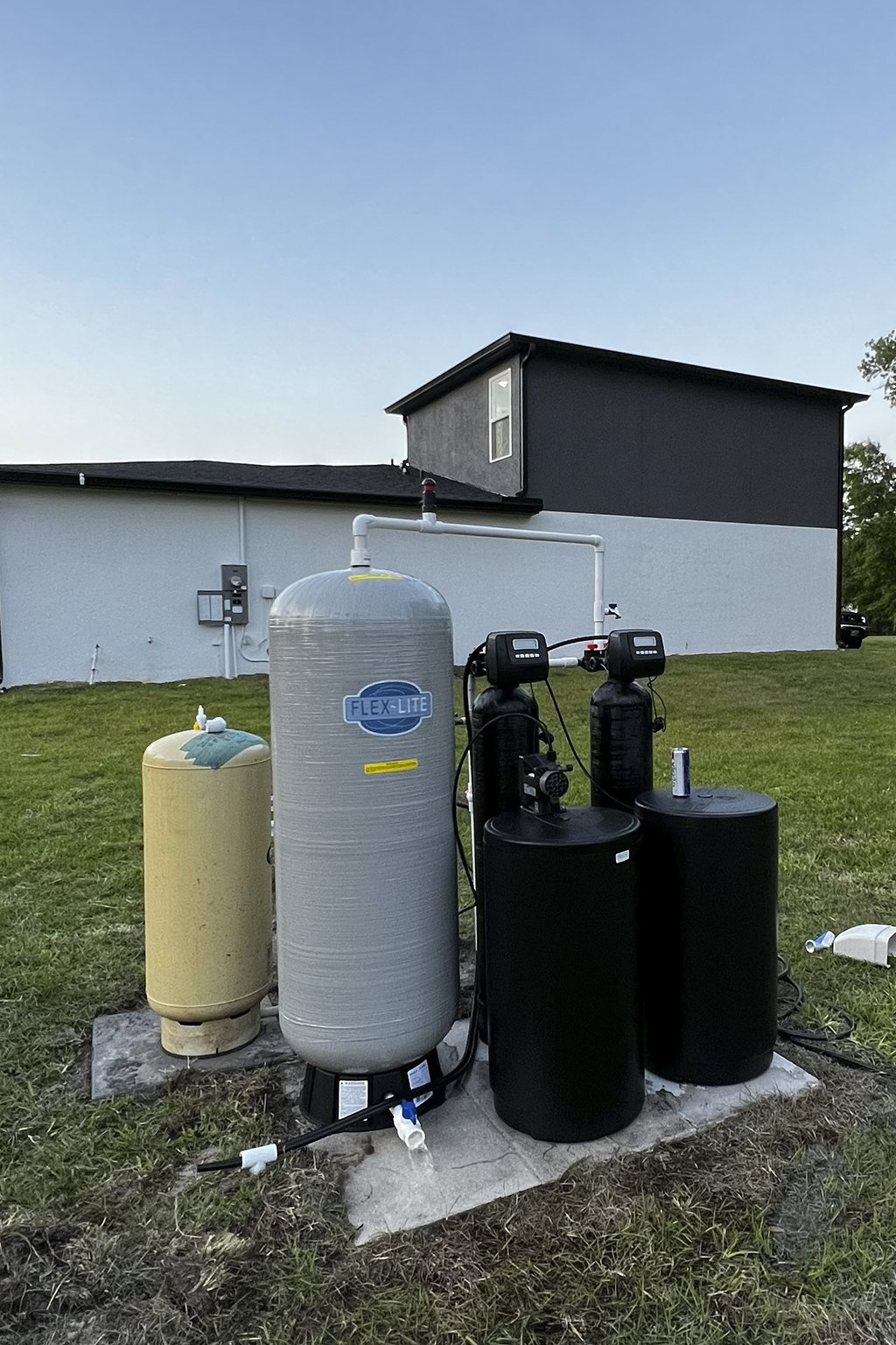 Outdoor water treatment system with large gray filter tank labeled 'FLEX-LITE,' two black control units, a yellow tank, and piping set on a concrete pad in a grassy yard near a white and black building.