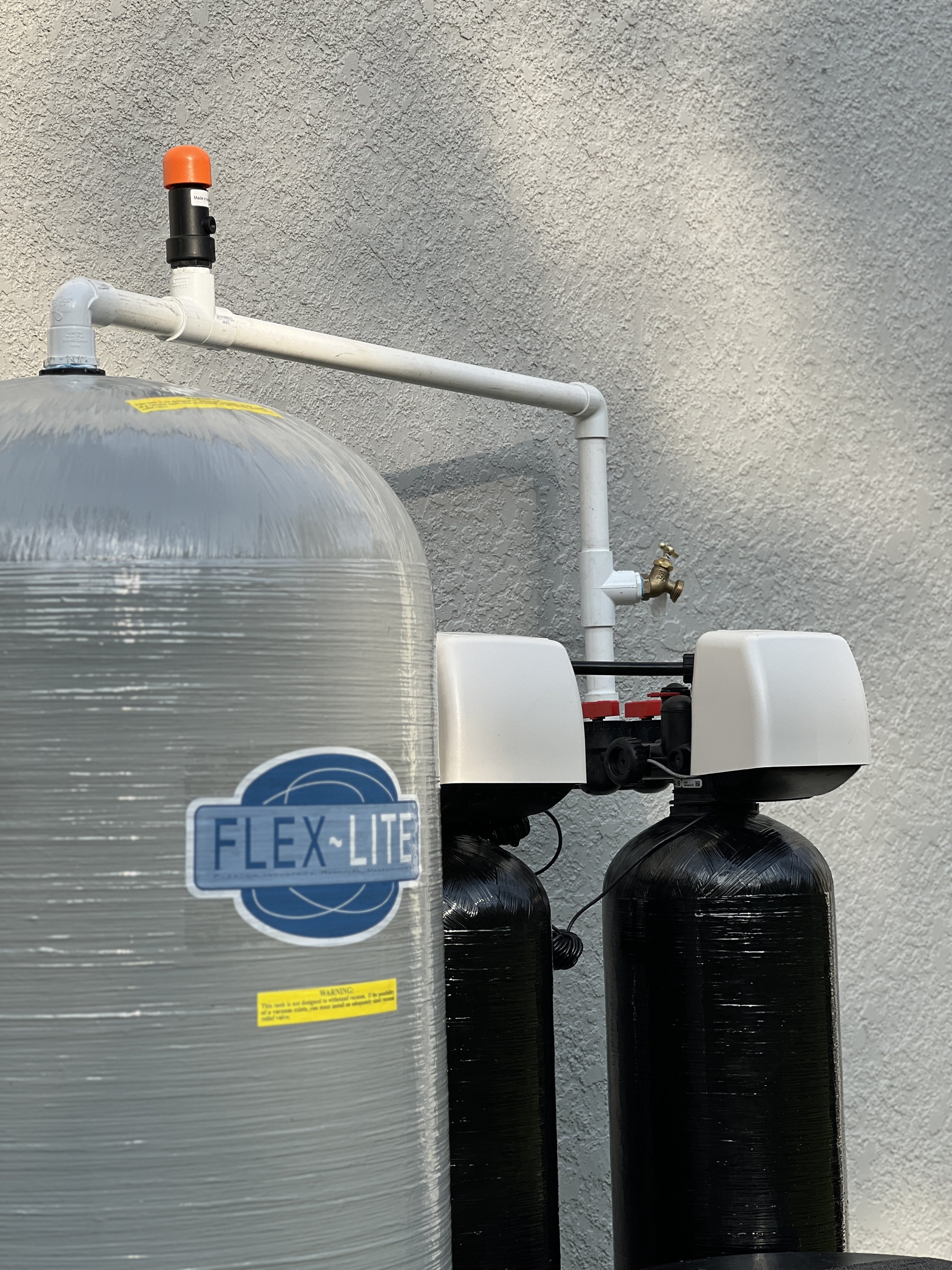 Close-up of water filtration system with a large clear and black tanks connected by white pipes against a textured wall.