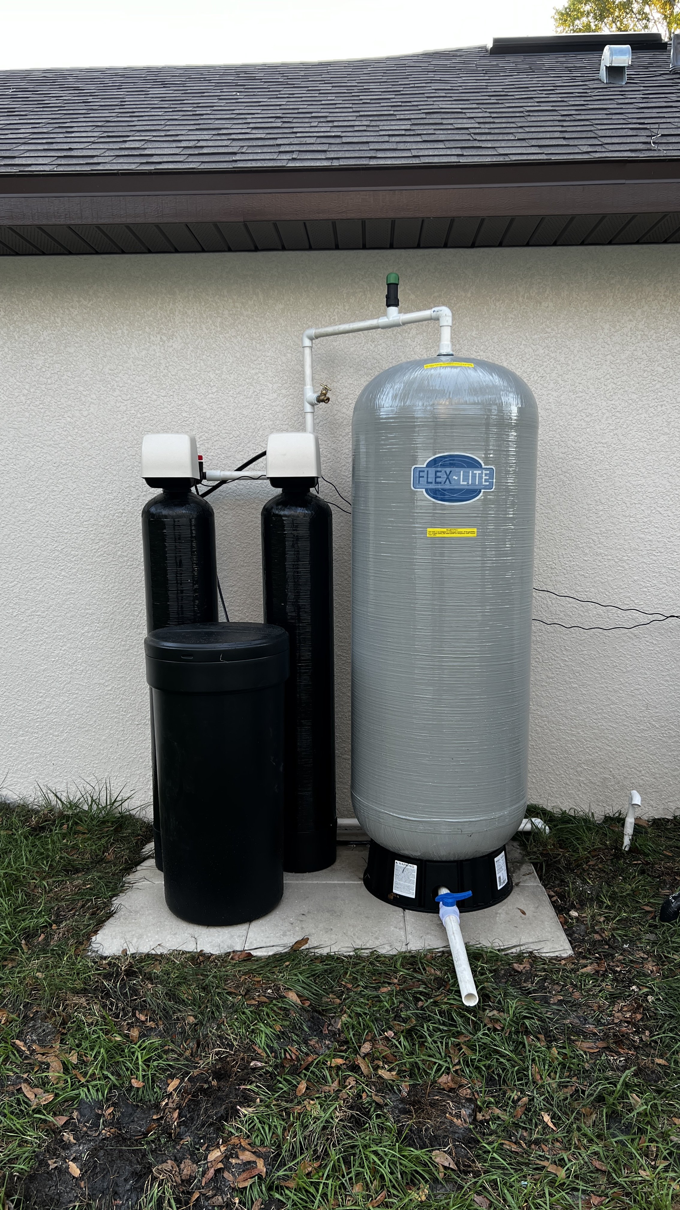 Outdoor water filtration and softening system with a large transparent tank labeled 'FLEX-LITE', connected to black filtration units, installed against a house wall.