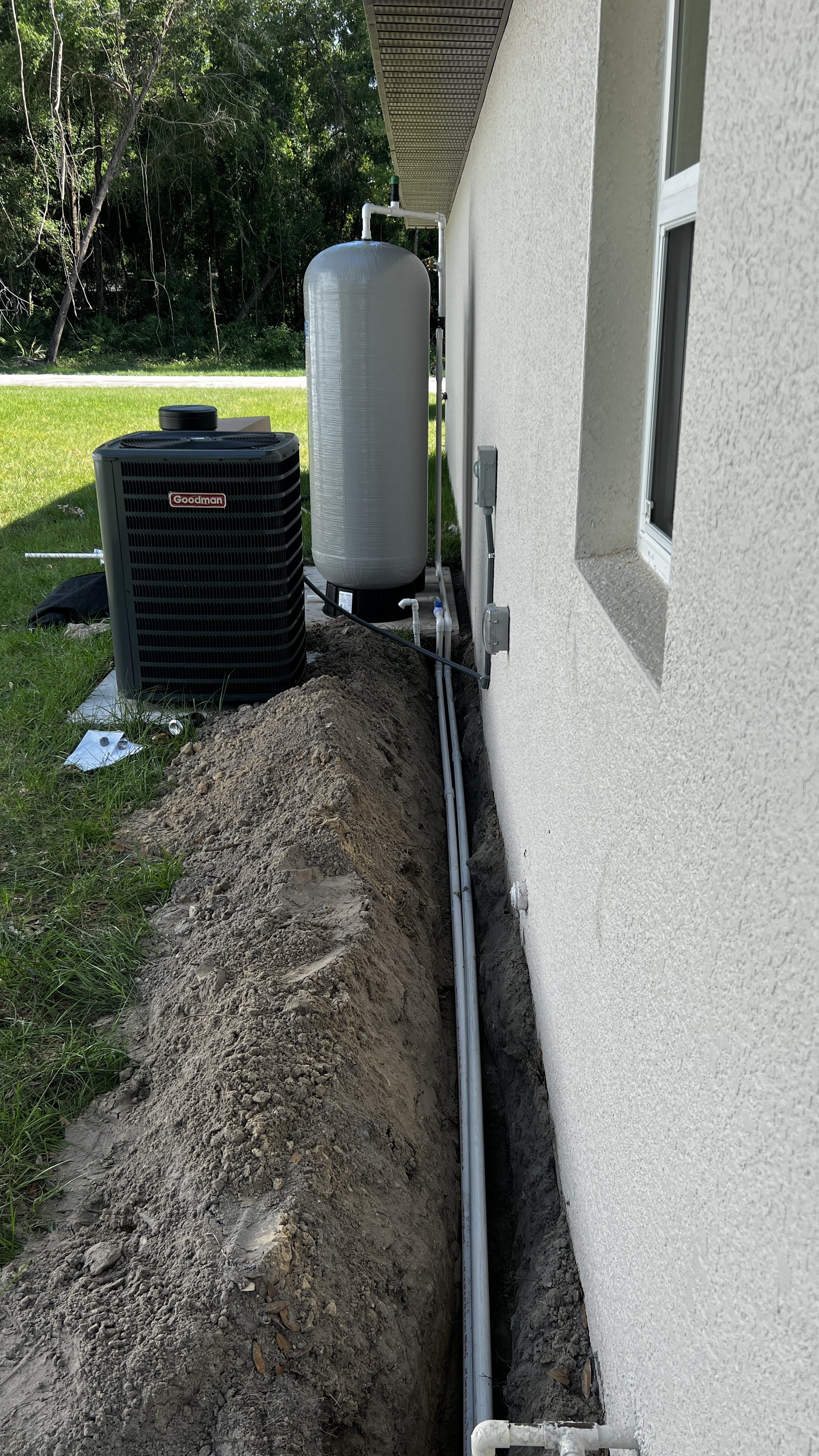 An outdoor view of a house's utility area showing an HVAC unit, a water filtration tank, and plumbing pipes installed near the house wall with a trench dug for the pipes.