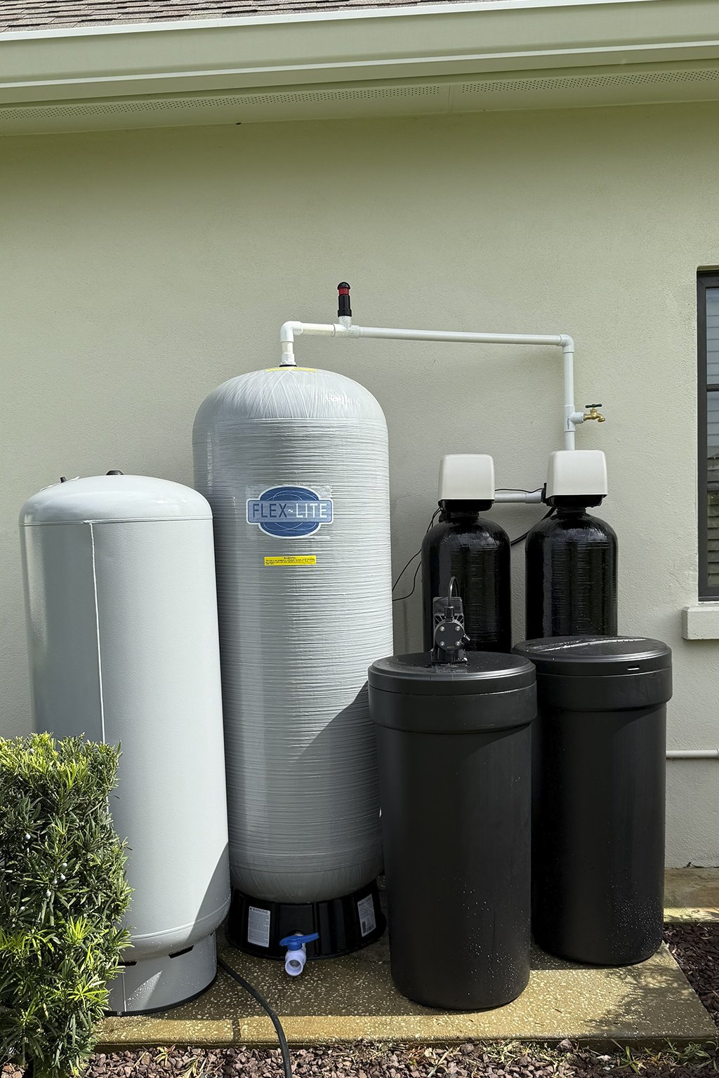 Exterior water treatment equipment including a large white tank, a gray pressure tank labeled 'FLEX-LITE,' and two black filtration tanks with control valves, situated beside a house wall with a window, on a concrete pad with a small shrub nearby.