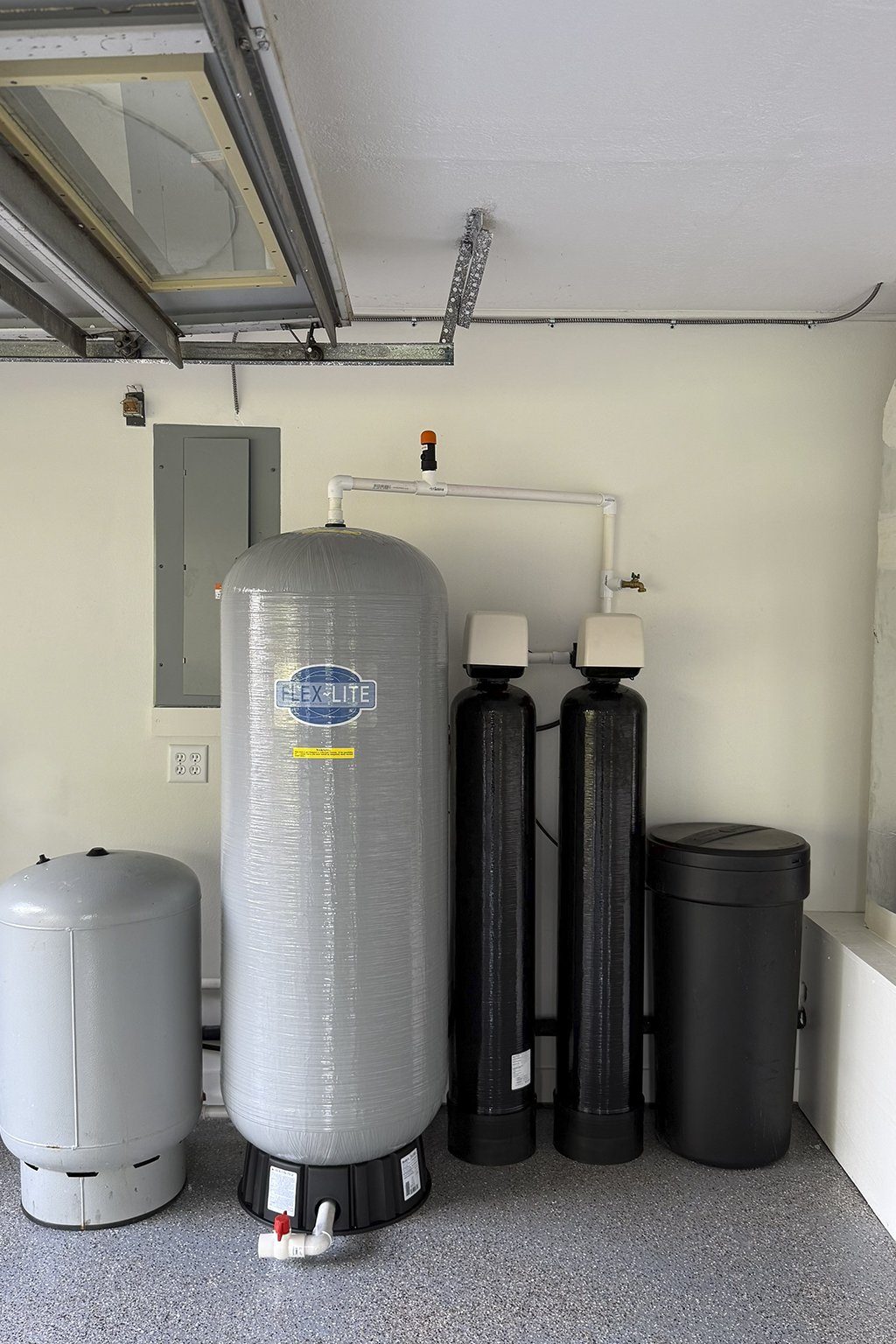 A water softener system with a large gray tank labeled 'Flexlite' and two black resin tanks with white tops, located in a utility room with a light-colored wall and gray speckled floor.