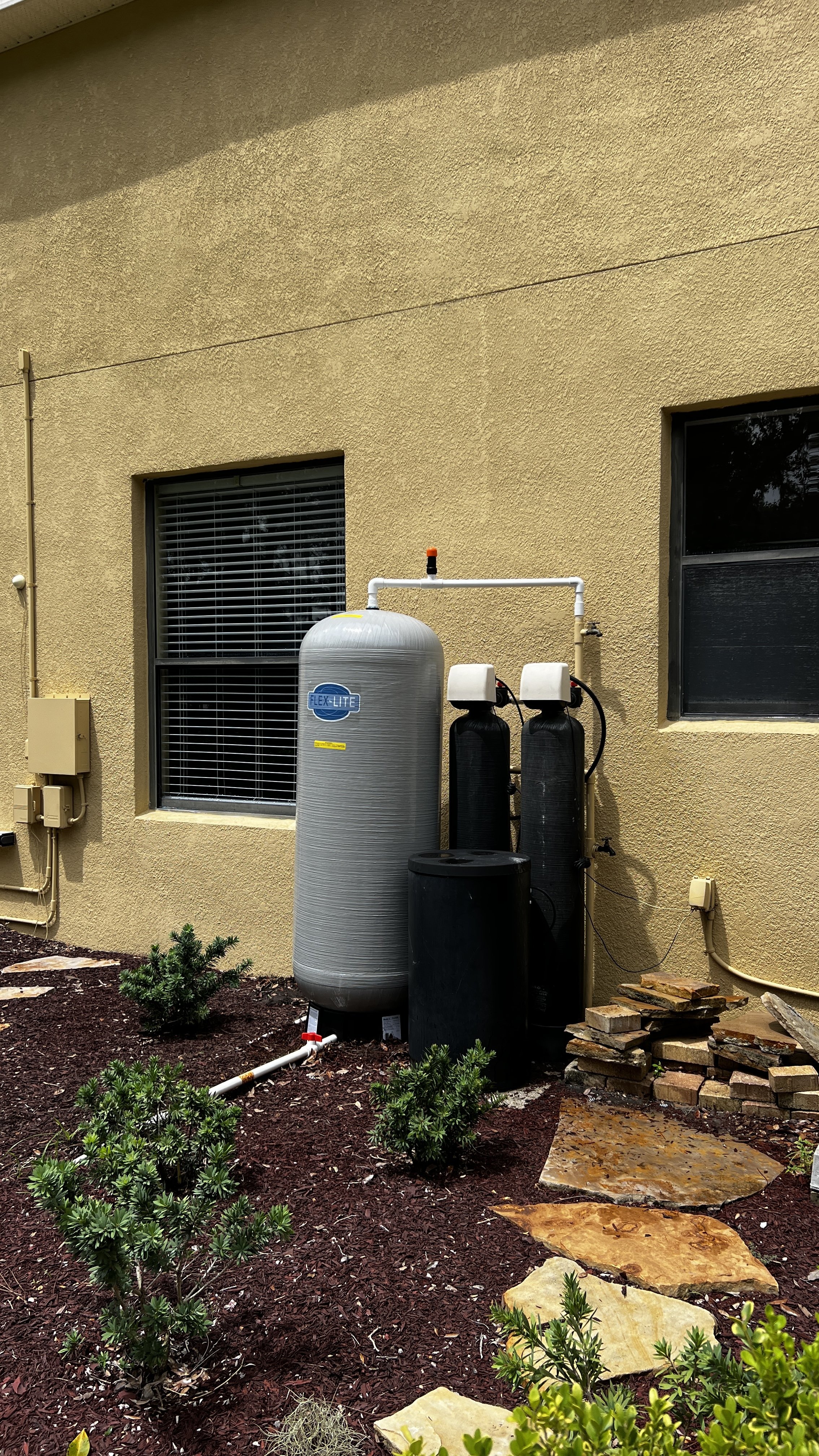 Outdoor water filtration system with large gray tank and black filters installed outside of a yellow building. Small green bushes and stone pathway in front.
