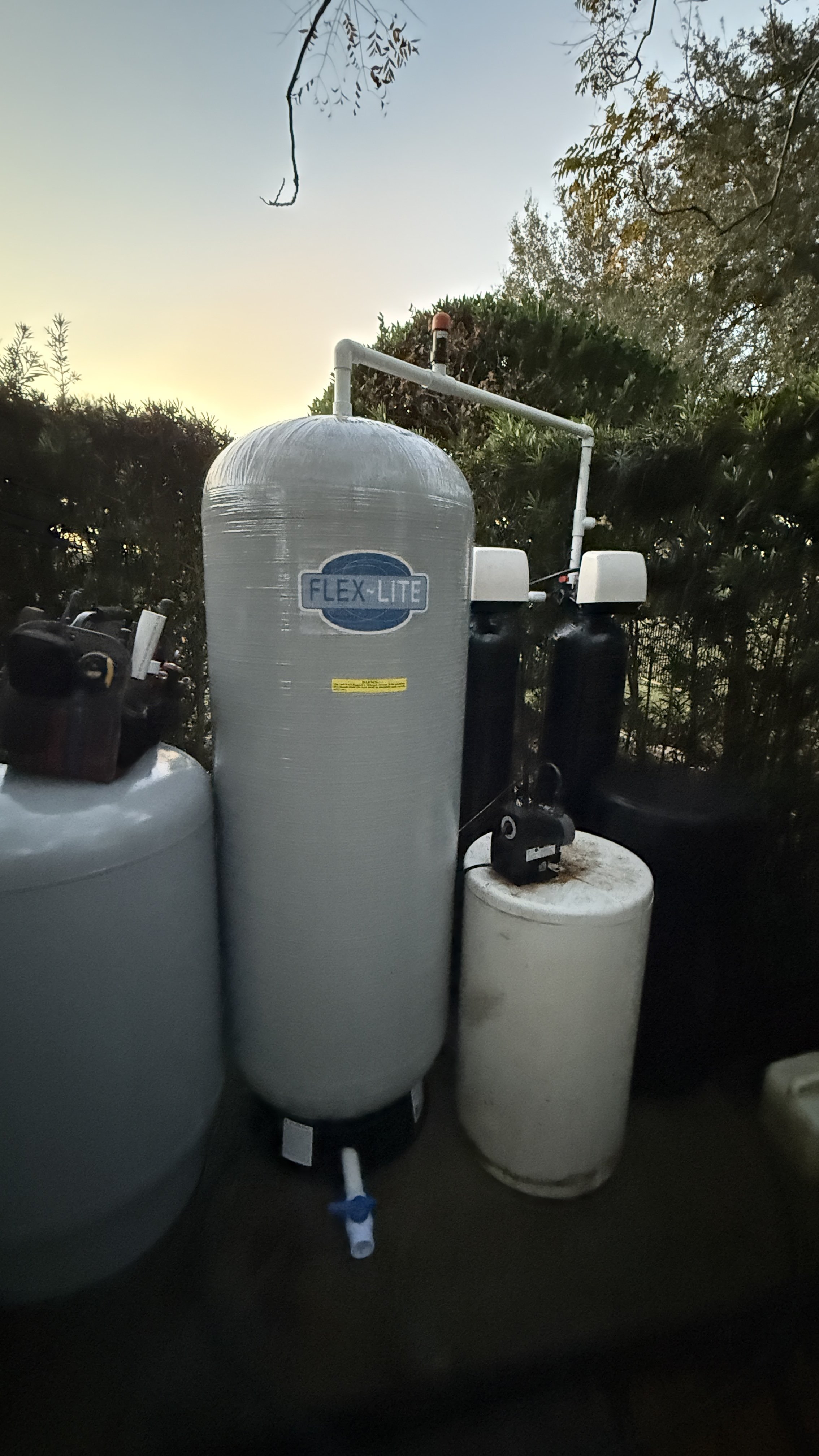 A water filtration system with a large gray tank labeled 'Flex-Lite' connected to pipes, black and white smaller tanks, and a gray cylinder, outdoors against a backdrop of trees and a dusk sky.