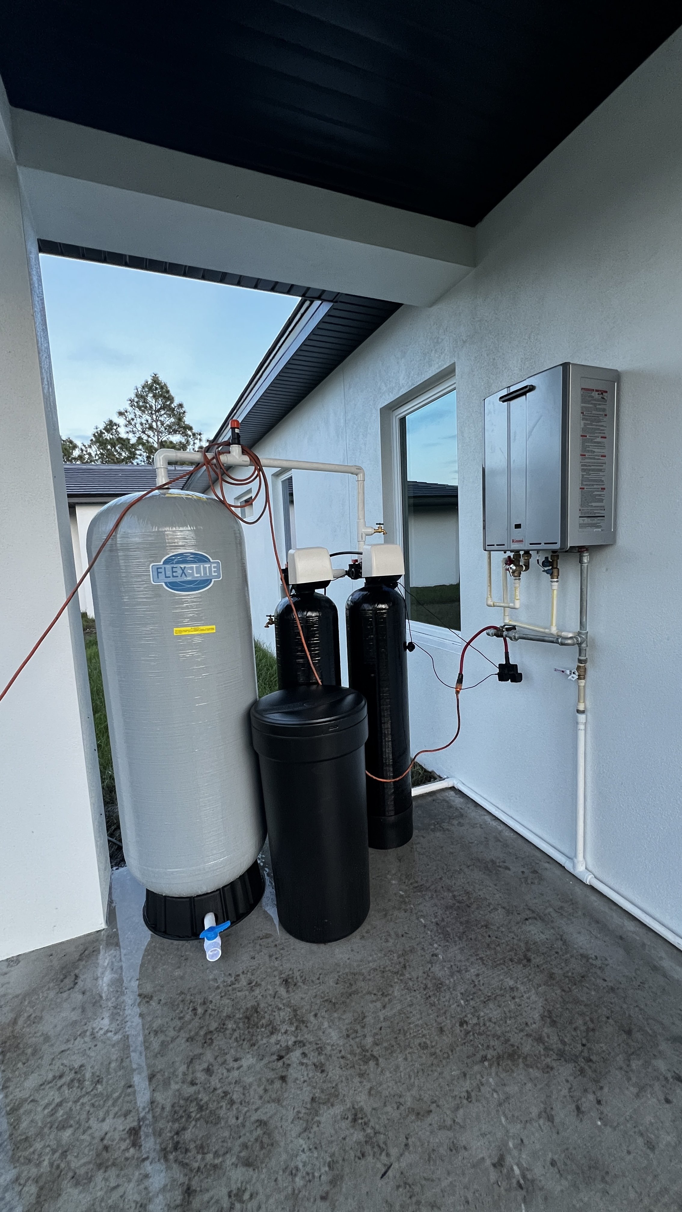 Outdoor water filtration system on the side of a house, with large white and black tanks, a control panel, and piping.