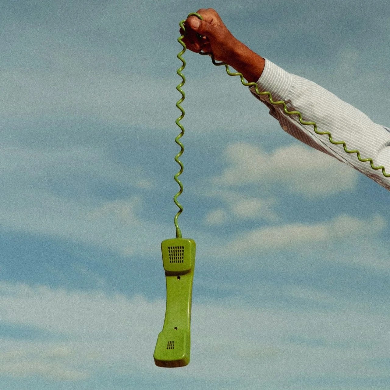 A person's hand holding a green corded vintage telephone against a cloudy sky.