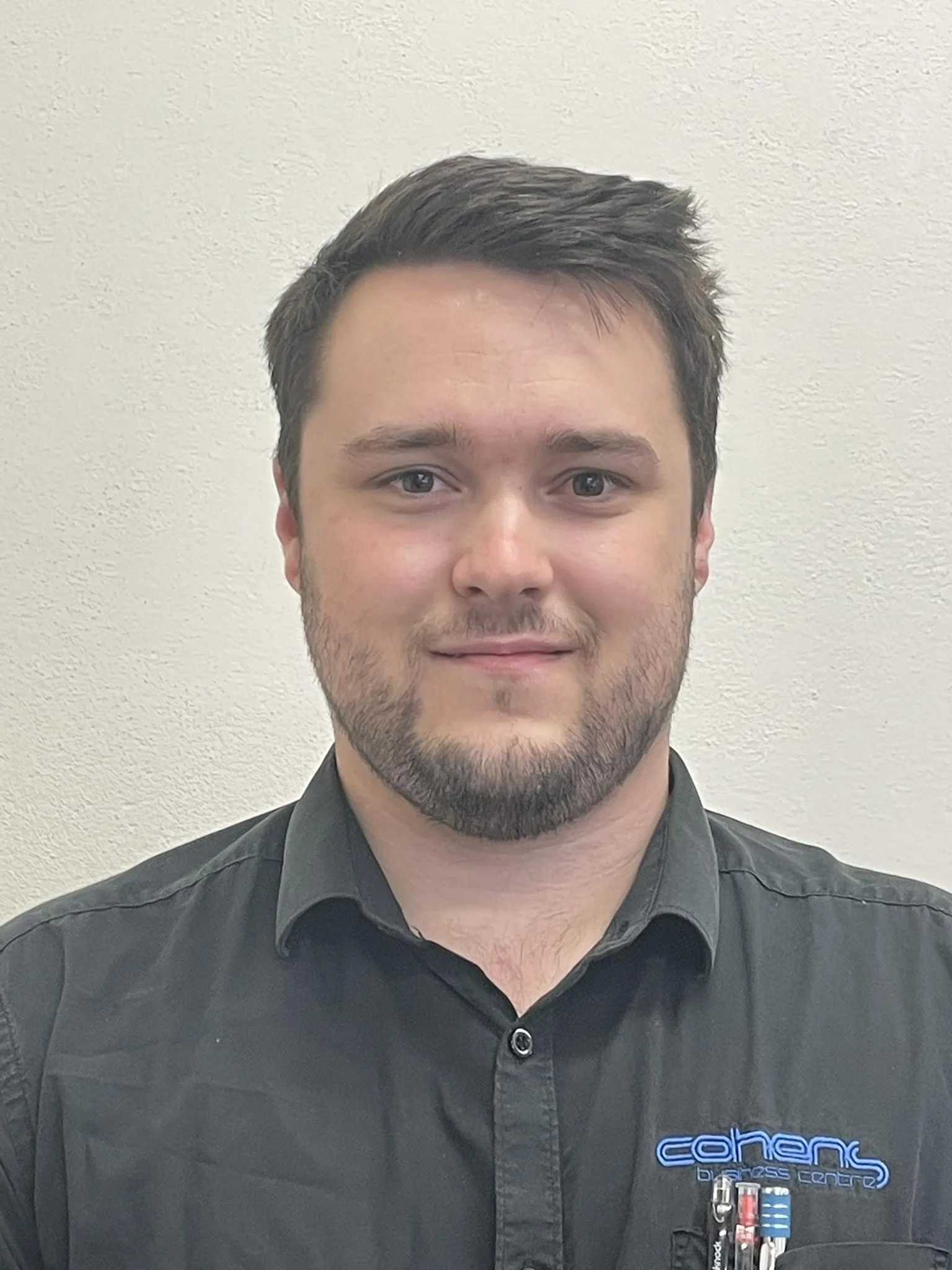 Headshot of a man with short brown hair, blue eyes, and a short beard, wearing a black collared shirt with a logo on it, standing against a plain light-colored wall.