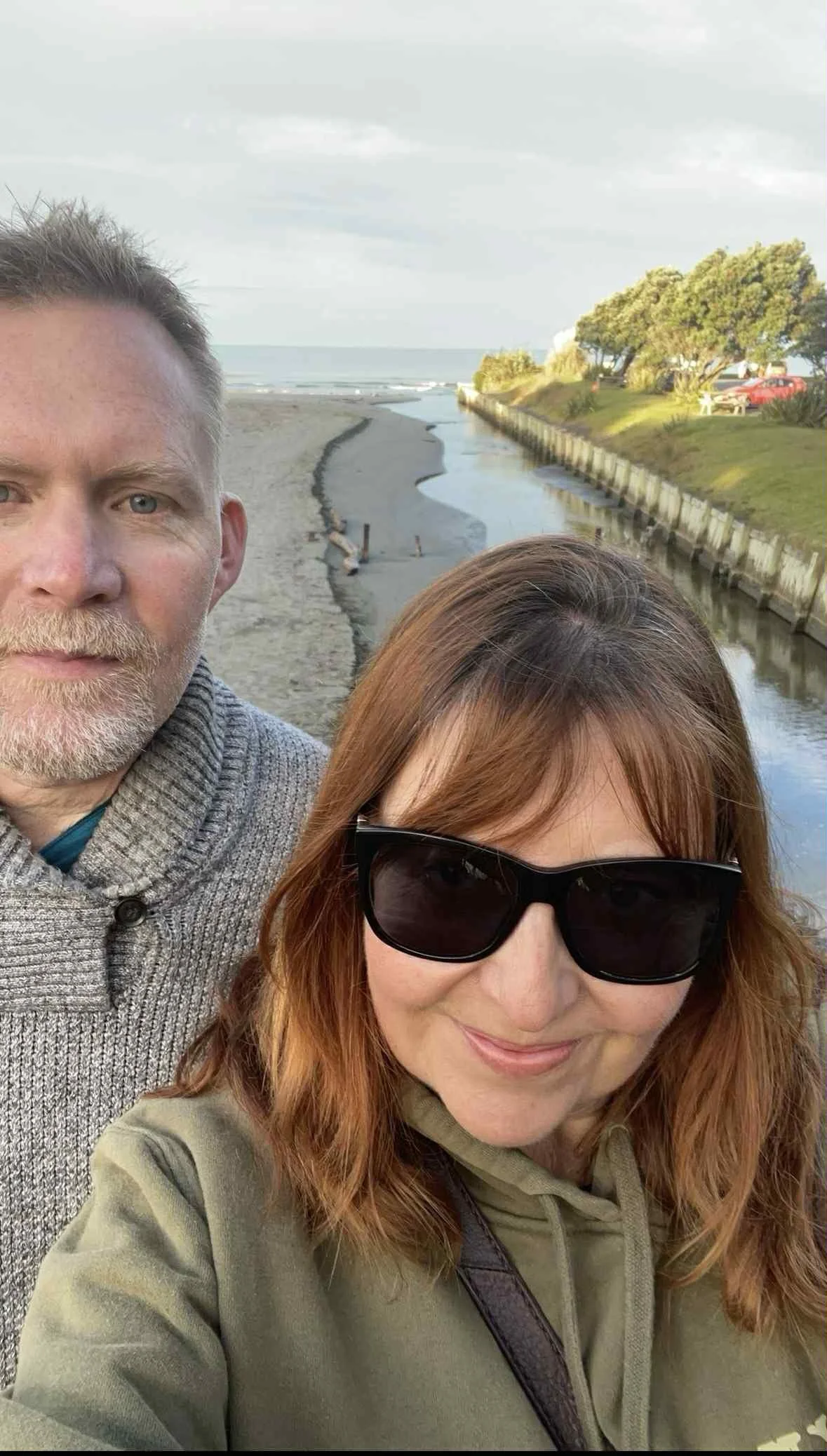 A woman with red hair, wearing sunglasses, smiling inside a glass gondola or observation wheel with a view of the landscape below.