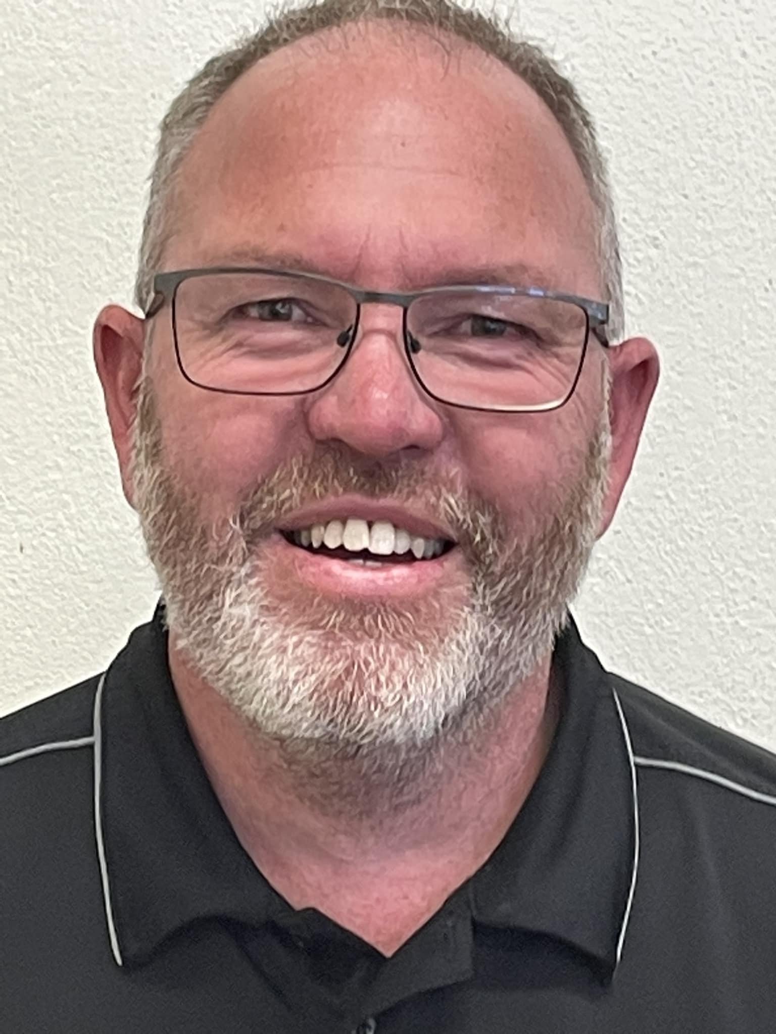 A smiling middle-aged man with glasses, a beard, and short hair, wearing a black shirt with white piping, standing against a plain, light-colored wall.
