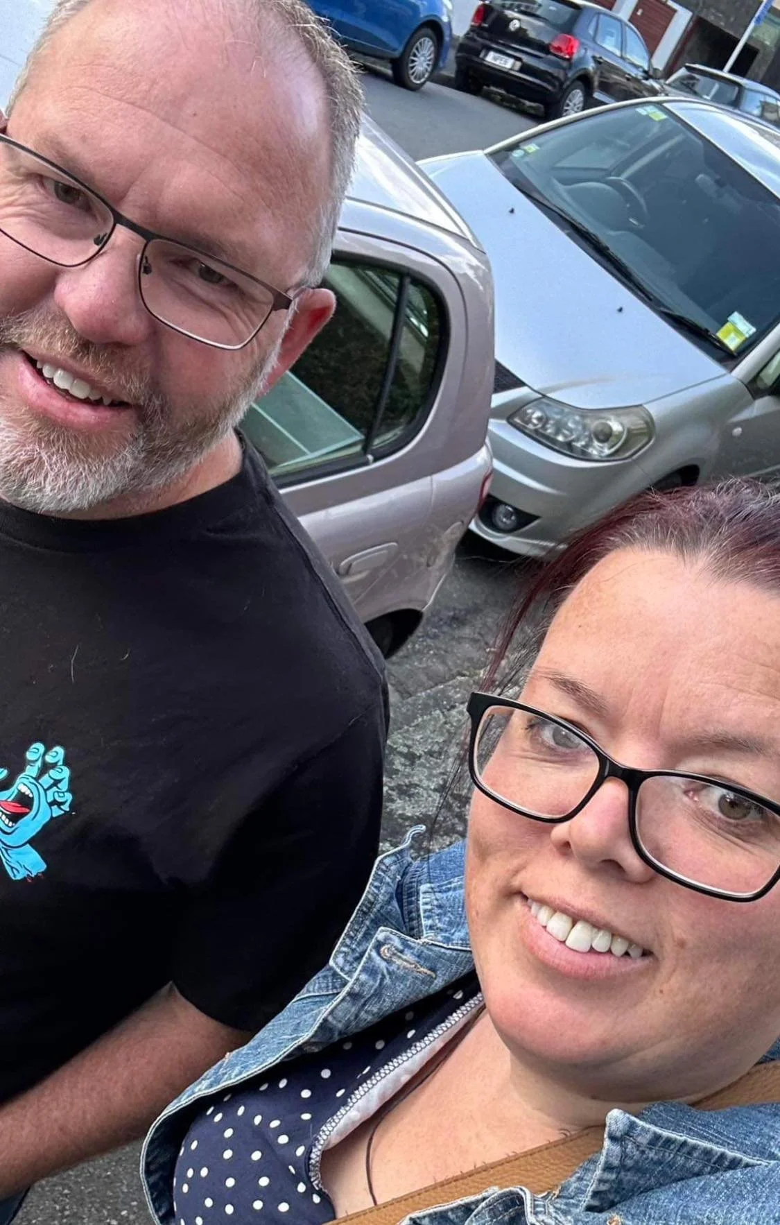 A selfie of a man and a woman outdoors, smiling at the camera, with parked cars in the background.