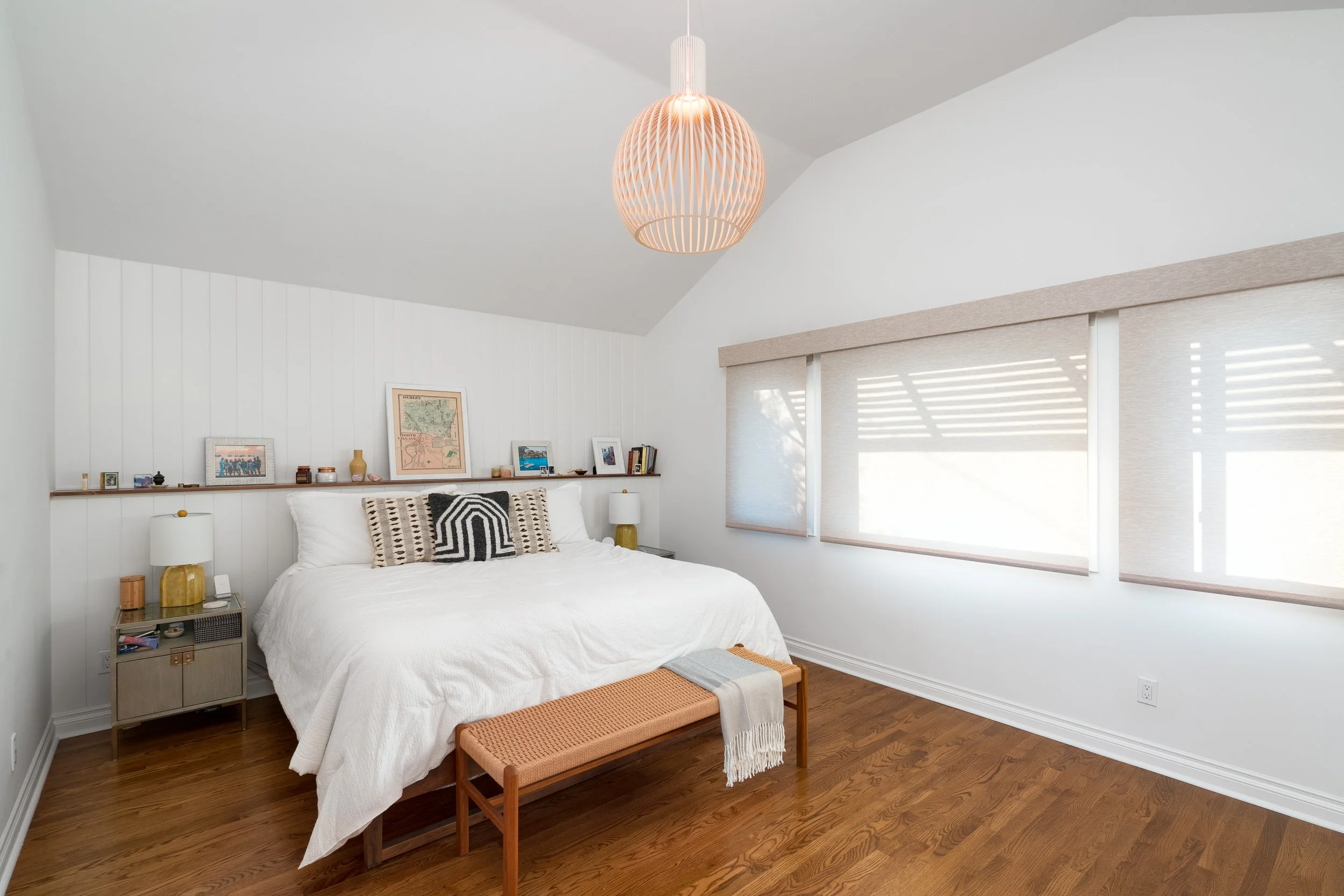 Minimalist bedroom with white bedding, wood bench, warm pendant lighting, and large window with natural light