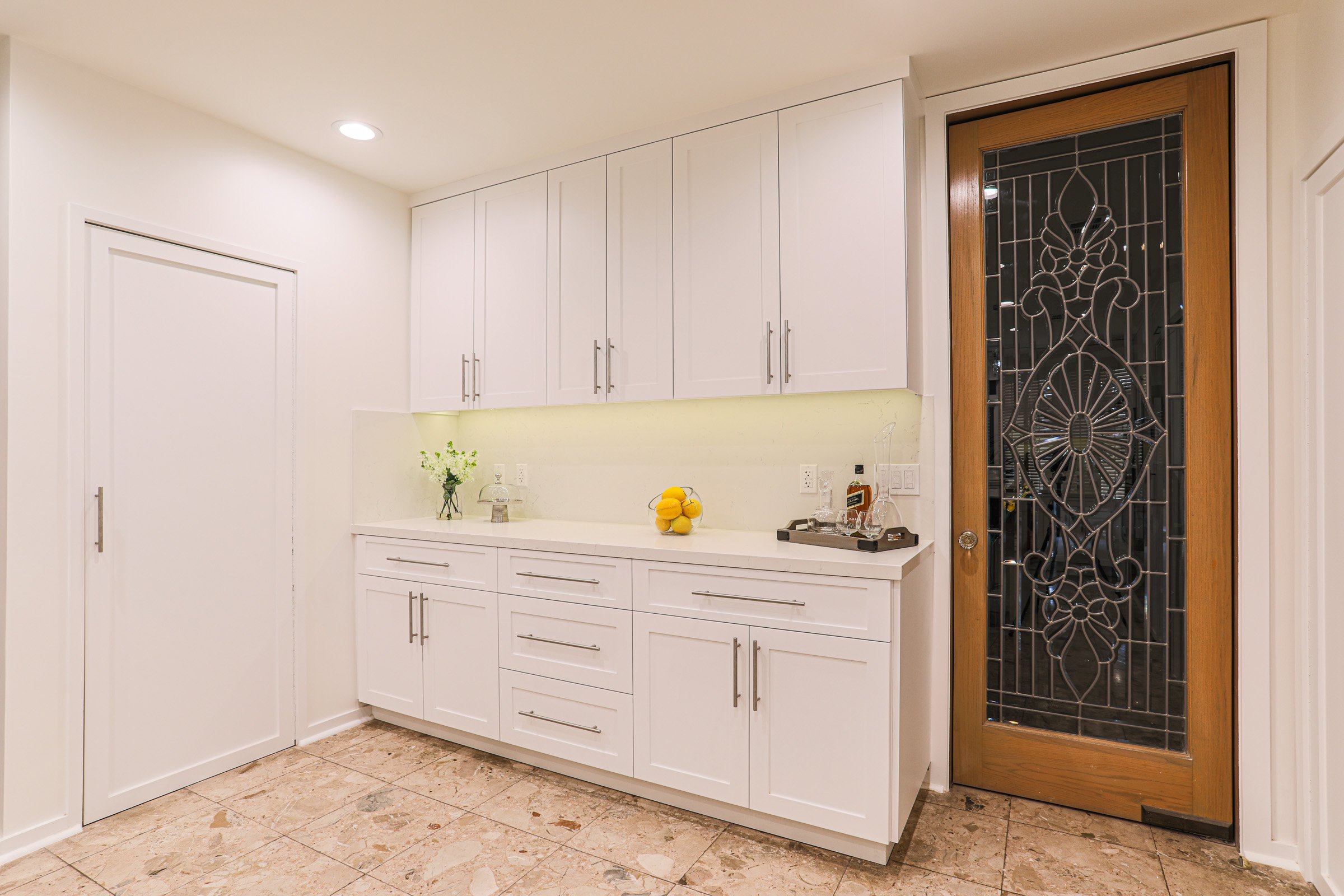 White cabinet kitchen with beige tiled floor, a door with a decorative metal grille, and a small bar cart with liquor bottles near the wall.