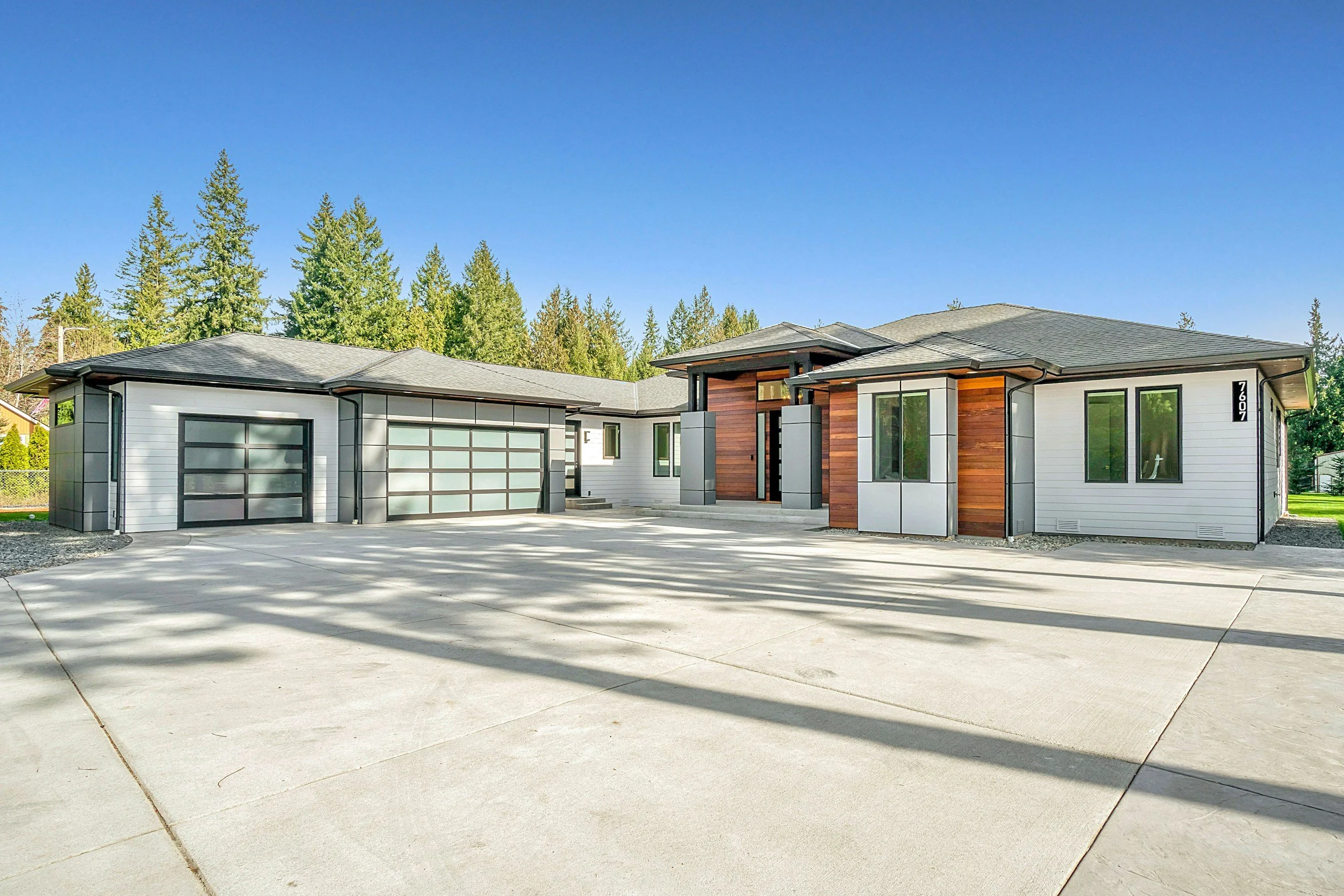 Modern single-family house with a concrete driveway, white and wood exterior siding, large windows, and a landscaped background with trees under a clear blue sky.