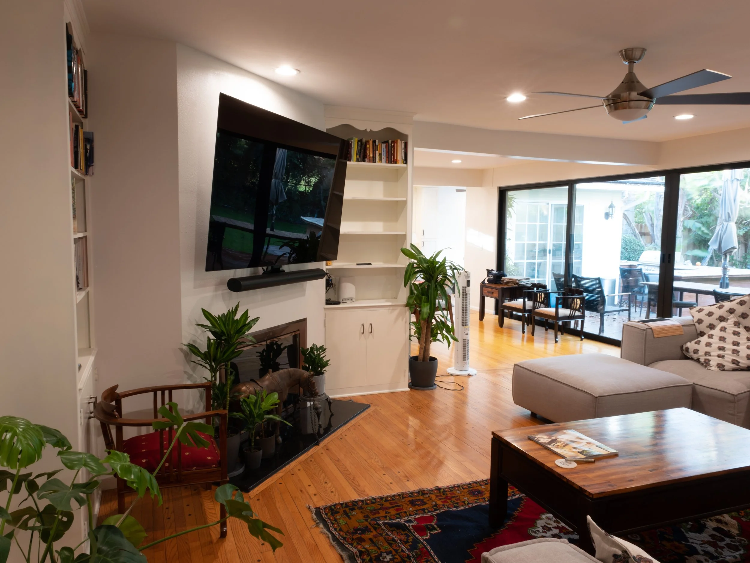 A living room with a mounted flat-screen TV above a fireplace, a beige sectional sofa, a wooden coffee table with magazines, and large sliding glass doors opening to an outdoor patio with chairs and umbrellas, decorated with potted plants and a colorful rug.