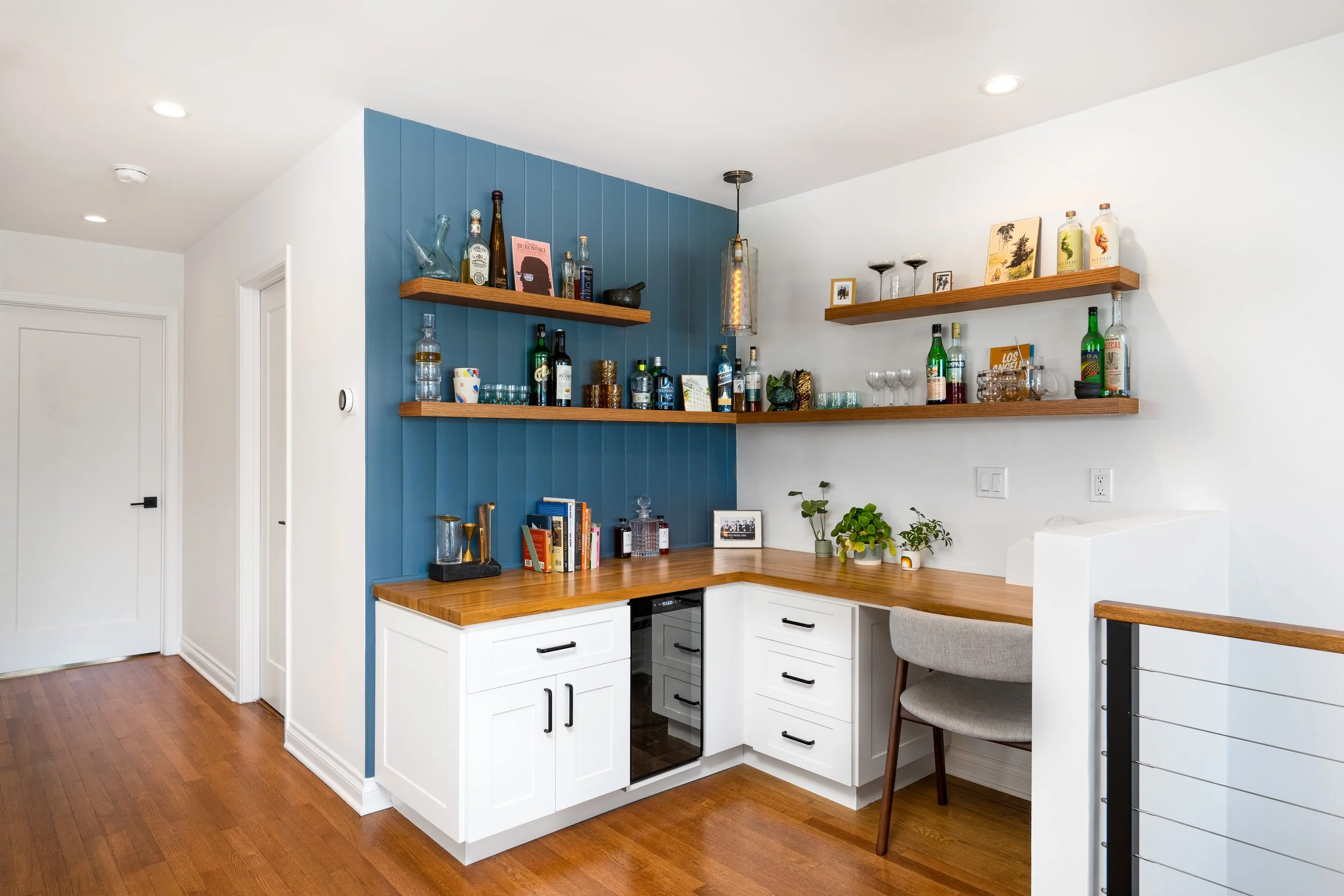 Home bar area with wooden countertops, white cabinets, blue feature wall with floating wooden shelves holding liquor bottles, glasses, and decor, a gray upholstered chair at the counter, and wooden flooring.