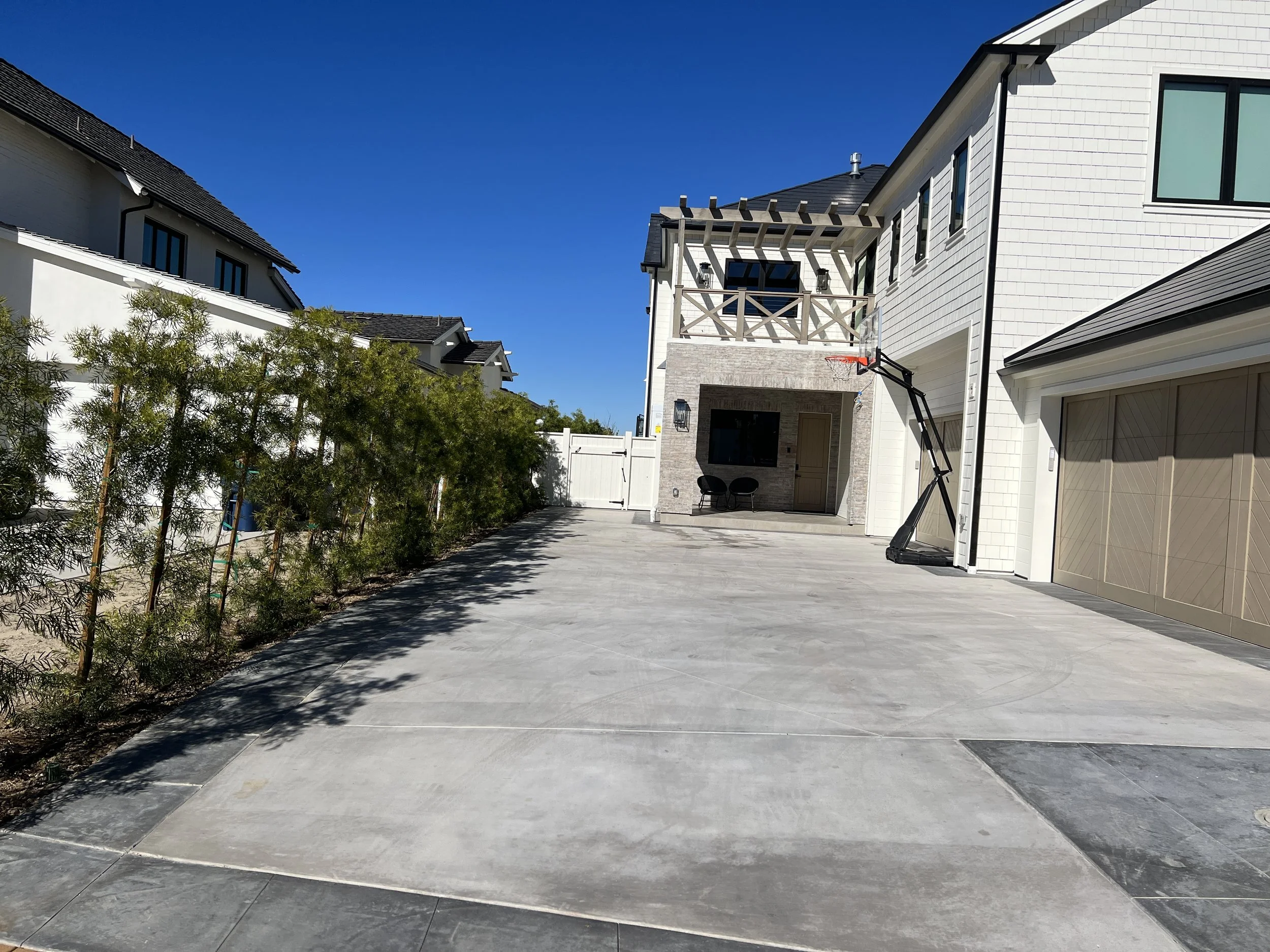 Spacious concrete driveway and side yard with clean landscaping, privacy fencing, basketball hoop, and access to a modern home exterior with balcony and outdoor living space.