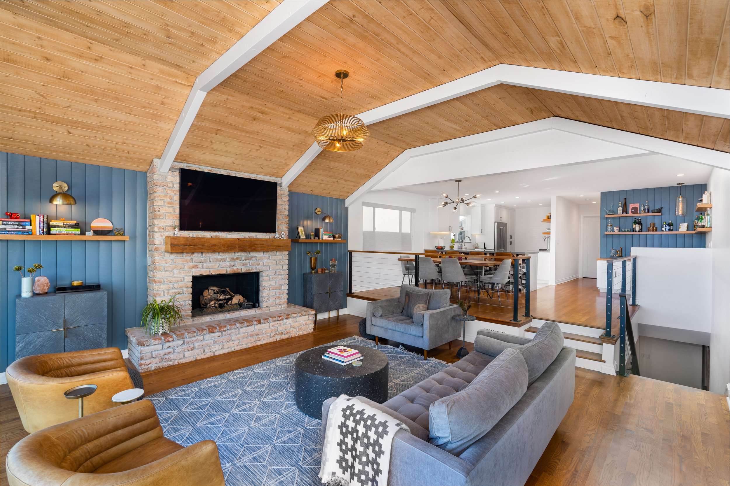 Open-concept living room with vaulted wood ceiling, brick fireplace, blue accent walls, and elevated dining area with modern kitchen and built-in bar
