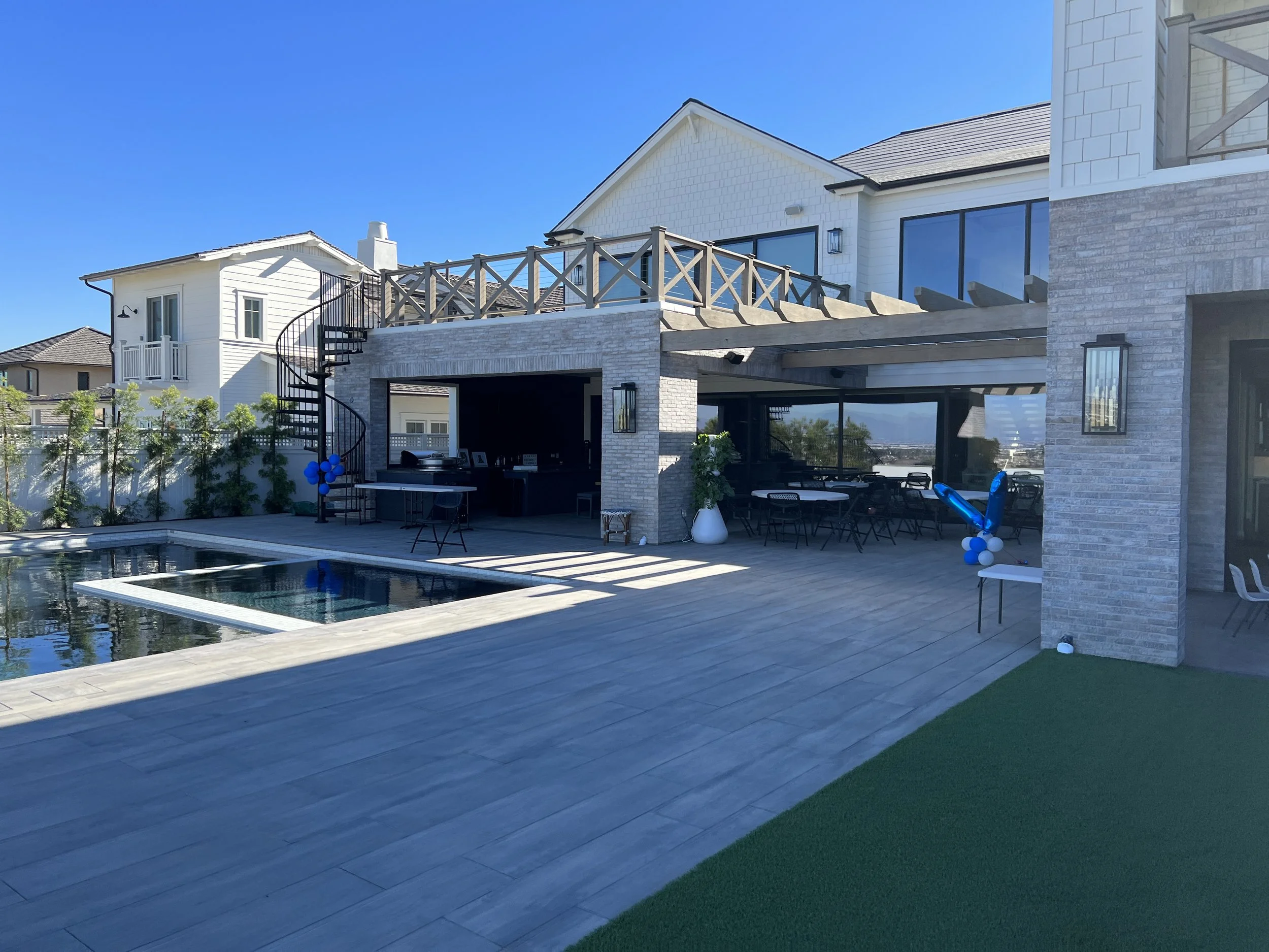 Modern backyard with a swimming pool, outdoor furniture, and a two-story house with a balcony, large windows, and outdoor lighting fixtures.