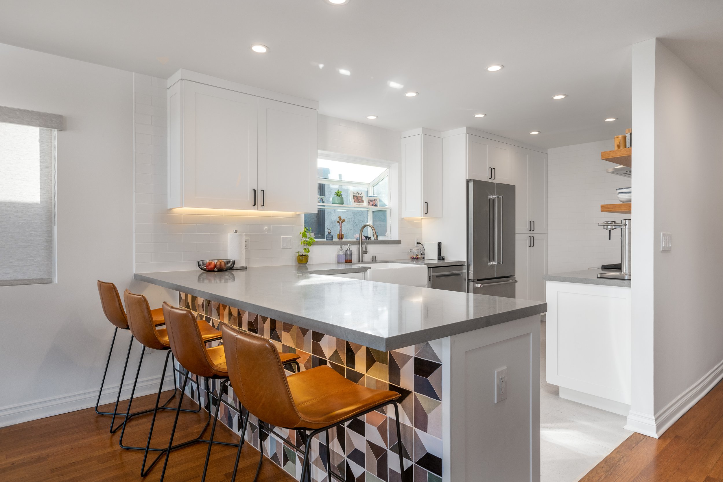 Modern kitchen with white cabinets, gray countertops, stainless steel refrigerator, sink under window, and four brown leather barstools at the counter with colorful geometric tile accents.