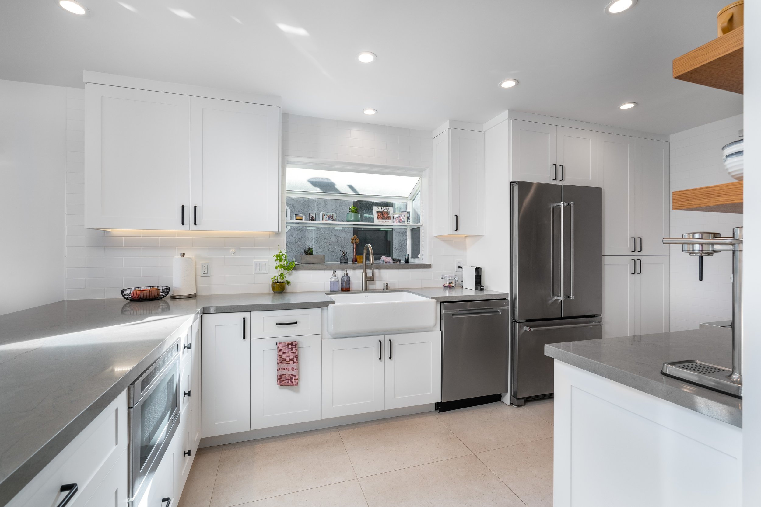 Modern kitchen with white cabinets, gray countertops, a white farmhouse sink, stainless steel refrigerator, microwave, and dishwasher, with a window above the sink and open wooden shelves on the right.