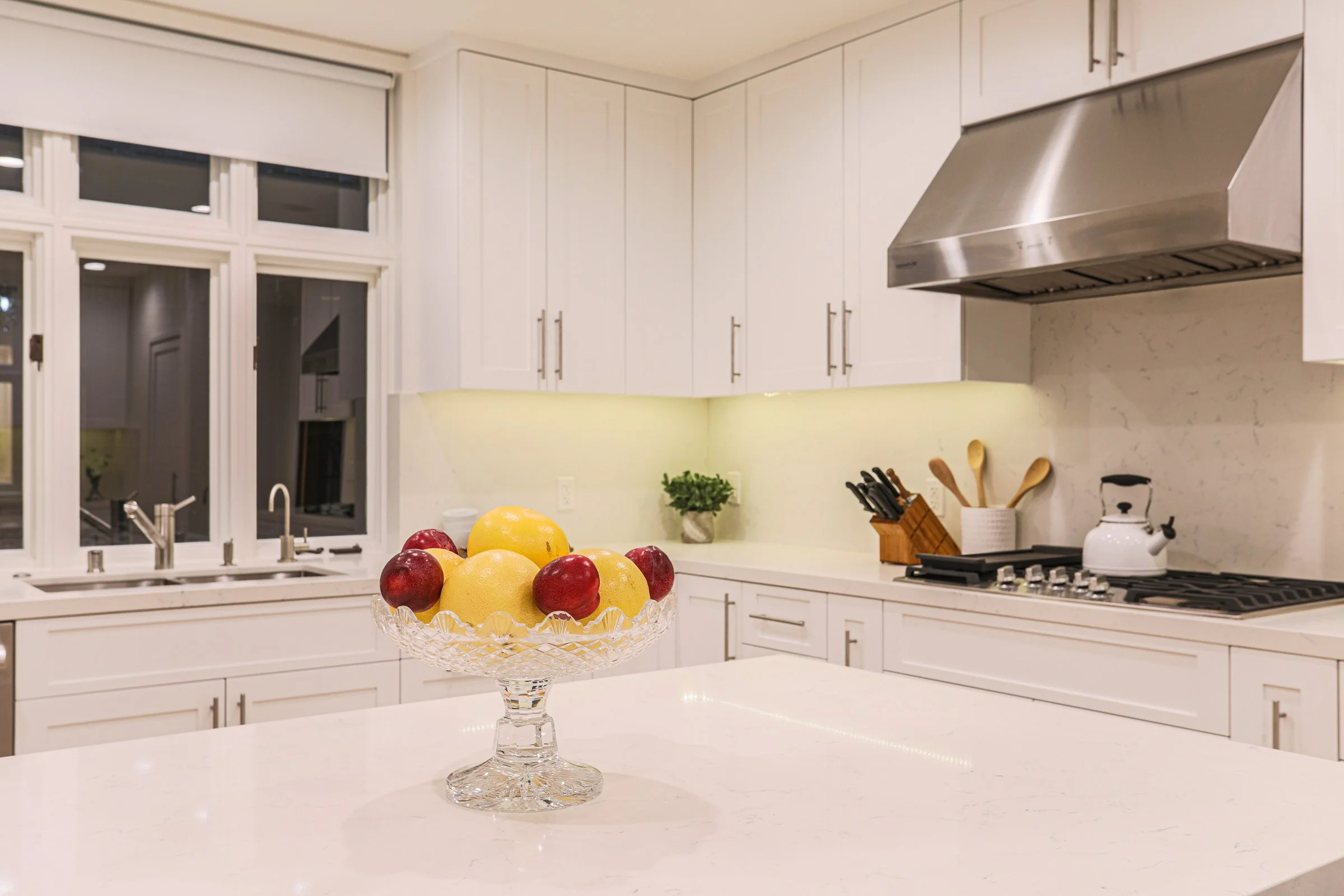 White kitchen with a central island, fruit bowl with lemons and apples, window, stove, kettle, and kitchen utensils.