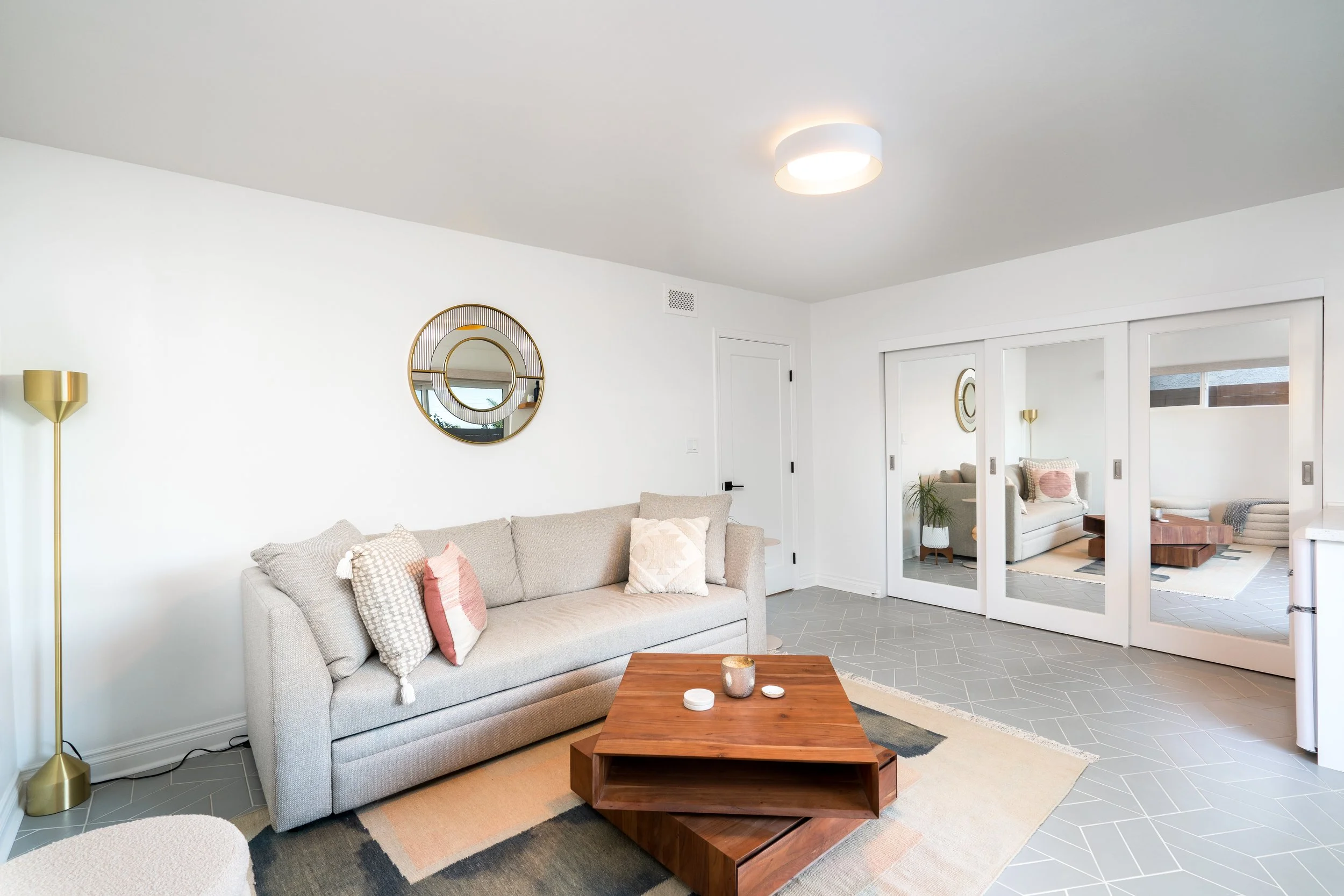 Modern living room with gray sofa, wooden coffee table, white walls, gold floor lamp, round mirror on wall, and glass sliding doors leading to another seating area.