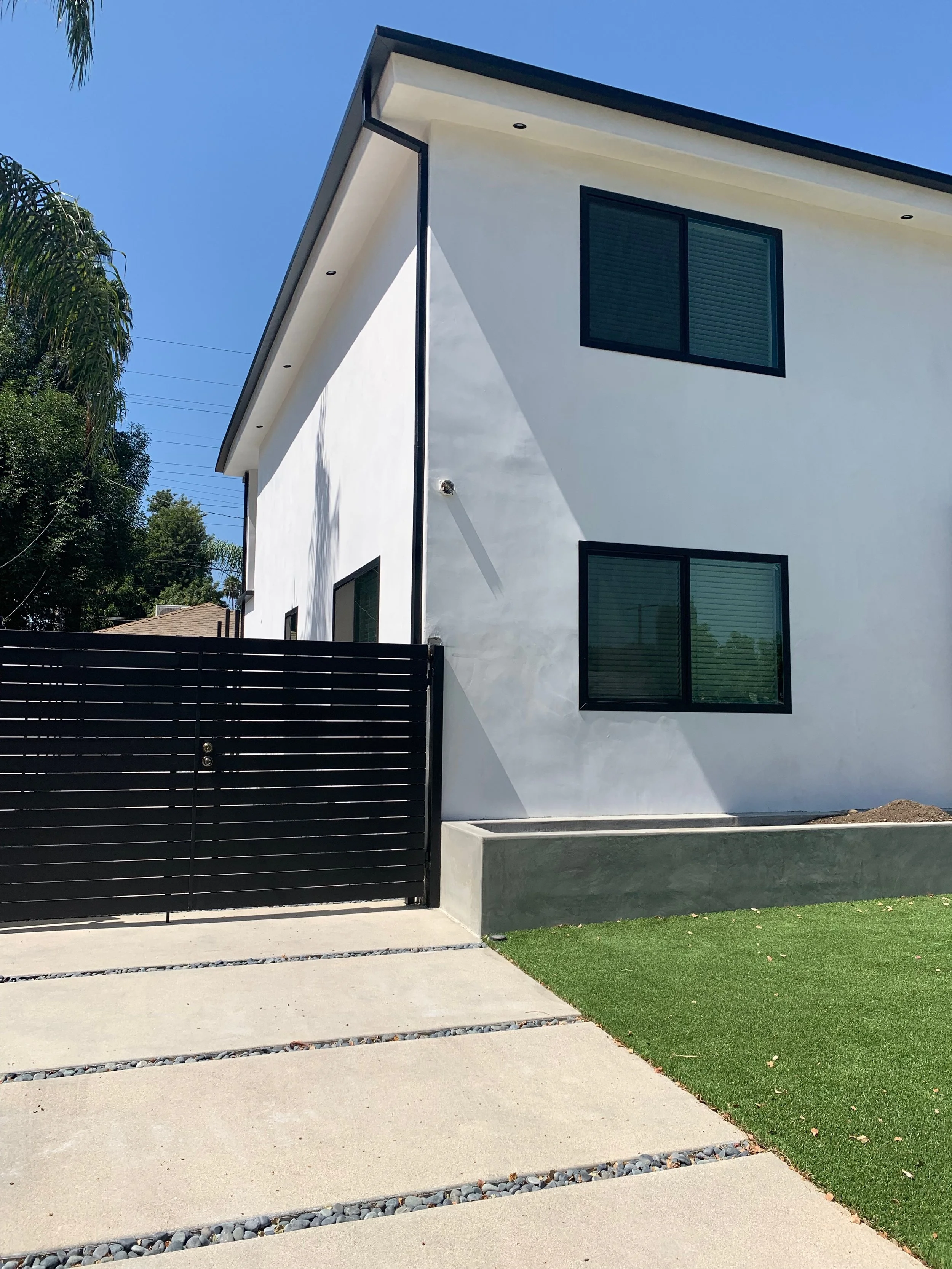 Modern white two-story house with black window frames, black fence, concrete driveway, and green lawn, under a blue sky.