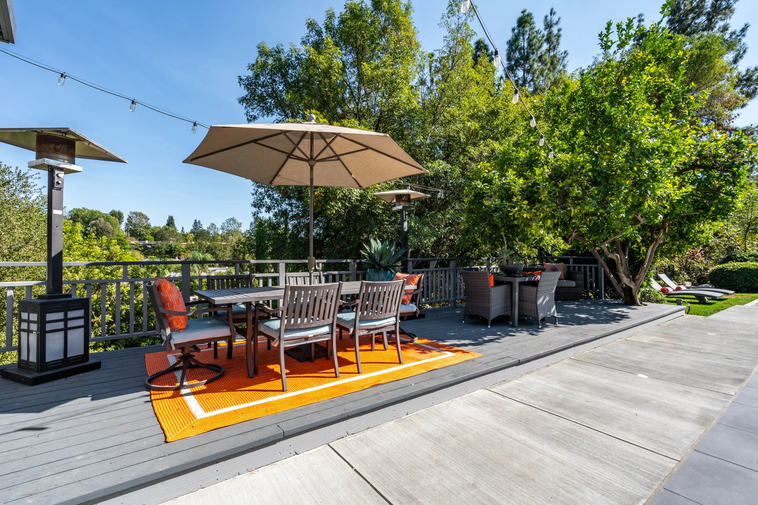 Outdoor patio with dining table, chairs, sun umbrellas, and lounge chairs surrounded by trees and greenery.