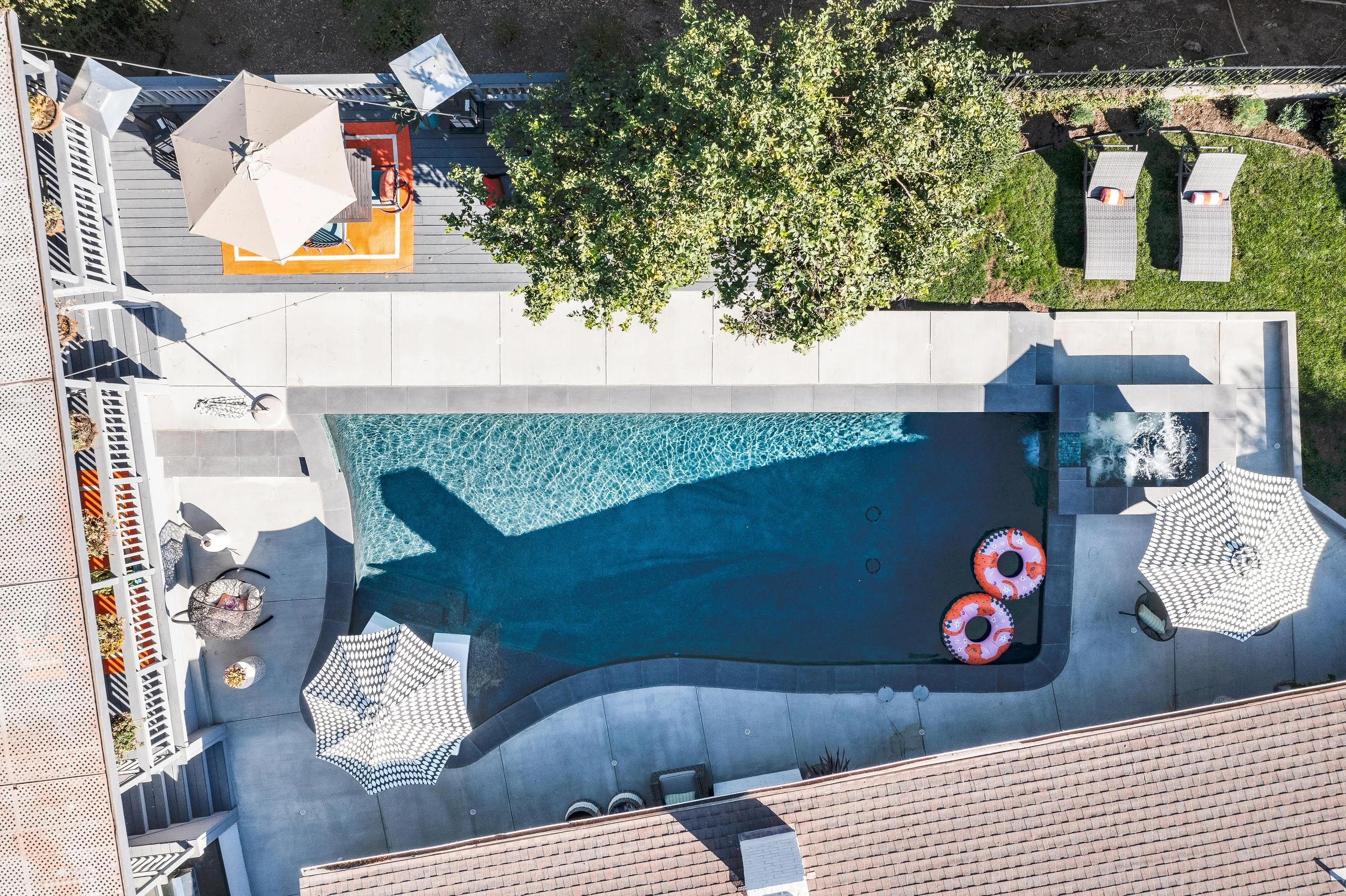Top-down view of a backyard with a swimming pool, lounge chairs, umbrellas, and a large tree.