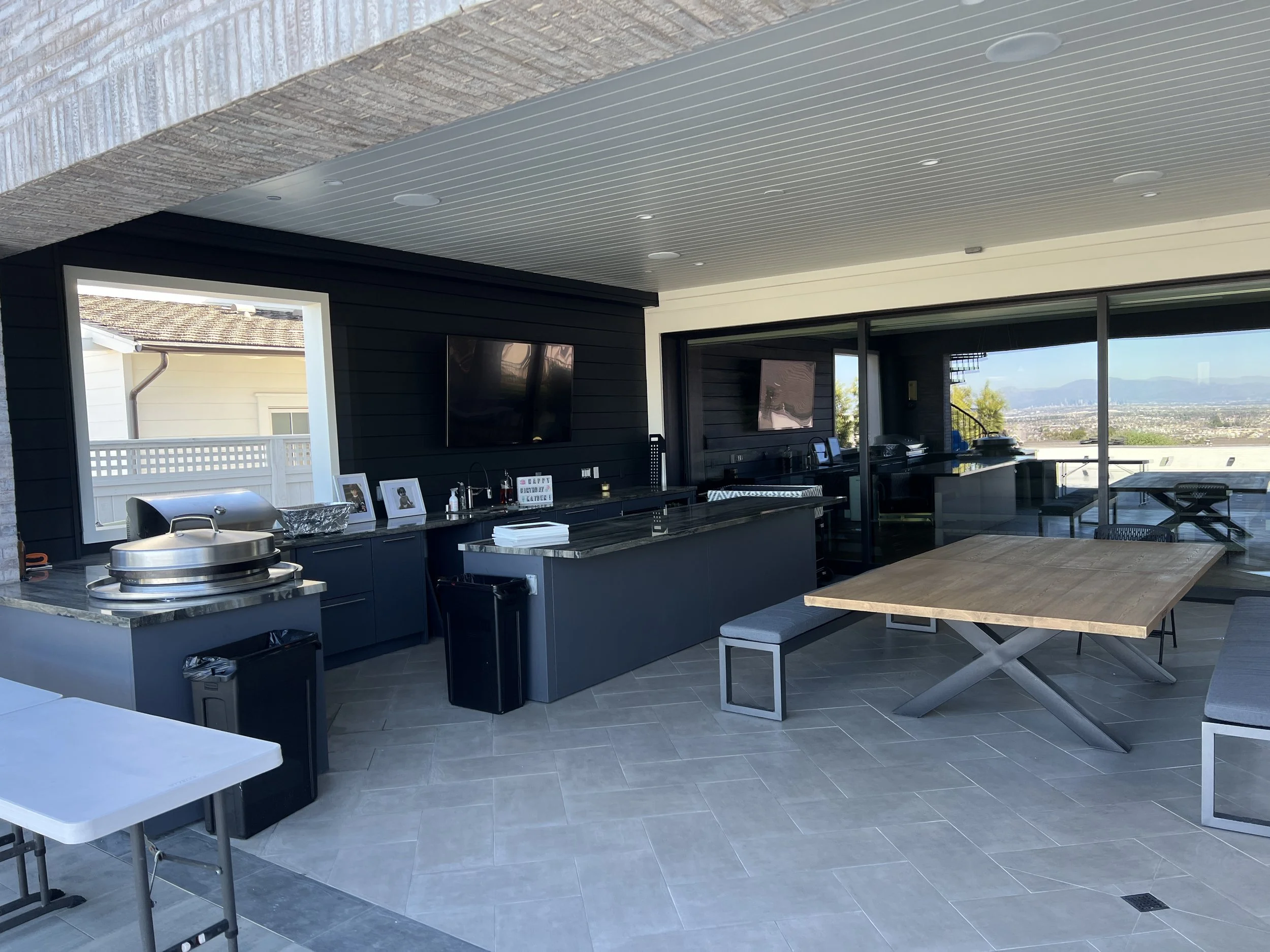 Covered outdoor kitchen and entertainment area with modern black cabinetry, built-in grill, wall-mounted TV, large sliding glass doors, and contemporary tile flooring.