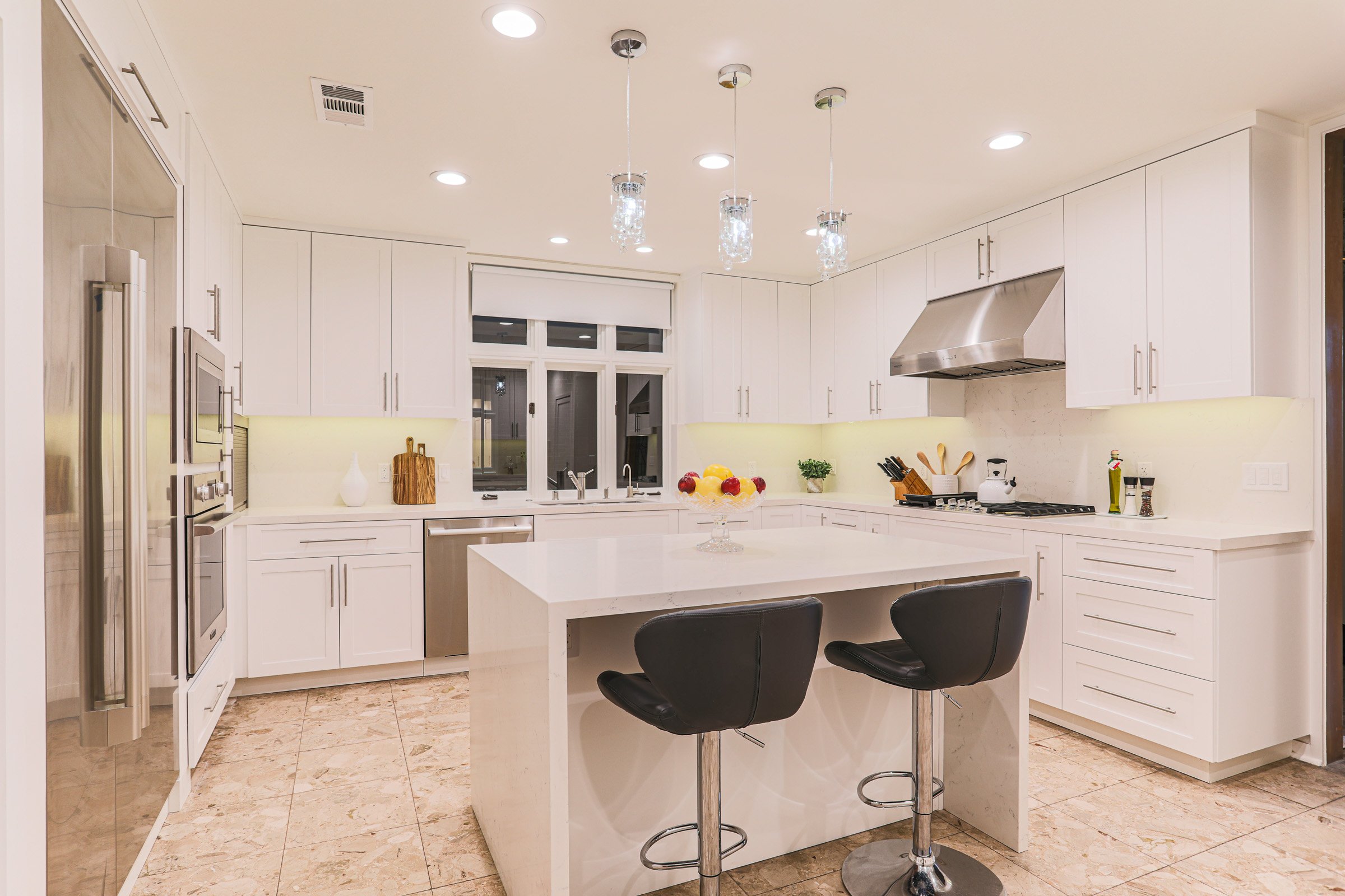 Modern white kitchen with pendant lights over an island, black chairs, built-in stainless steel appliances, and a bowl of fruit on the counter.
