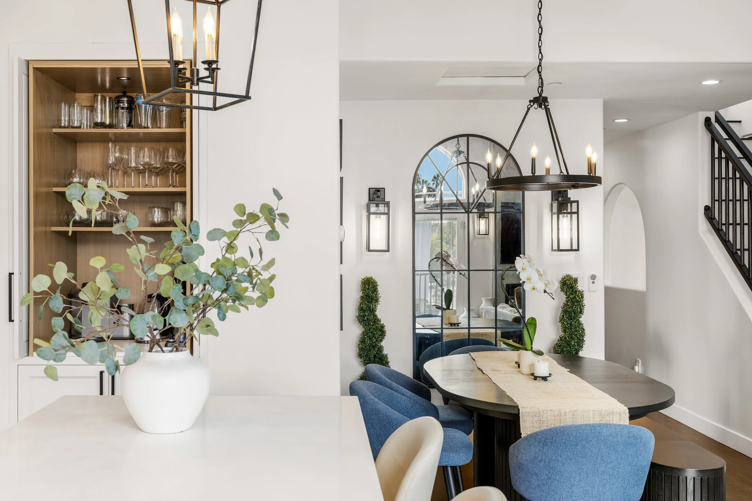 Custom dining area in a Manhattan Beach home remodel featuring built-in glassware storage, an arched black-framed mirror, statement chandelier, blue dining chairs, and modern black accents.
