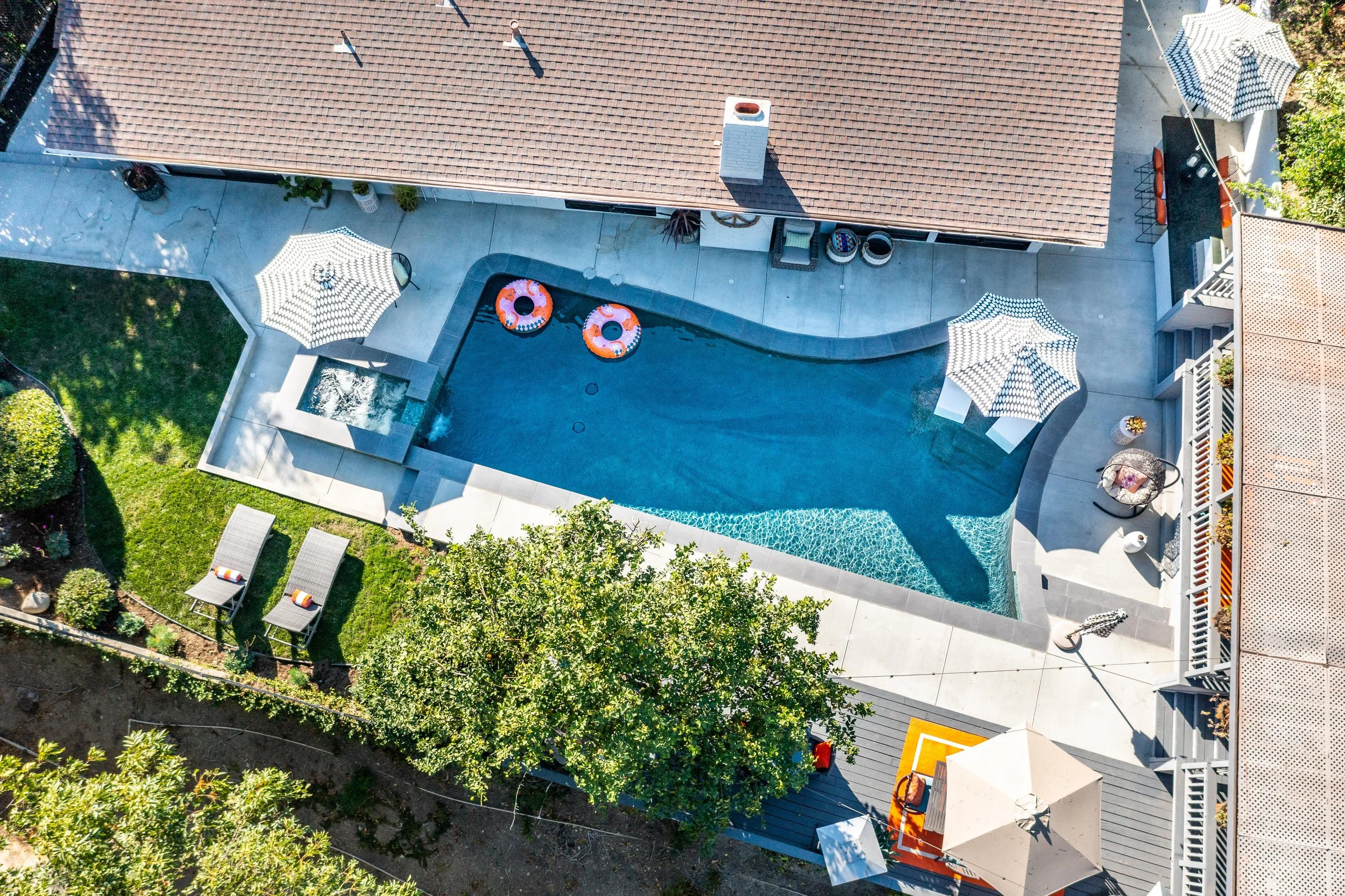 Aerial view of a backyard swimming pool surrounded by a concrete patio, patio umbrellas, lounge chairs, and greenery.