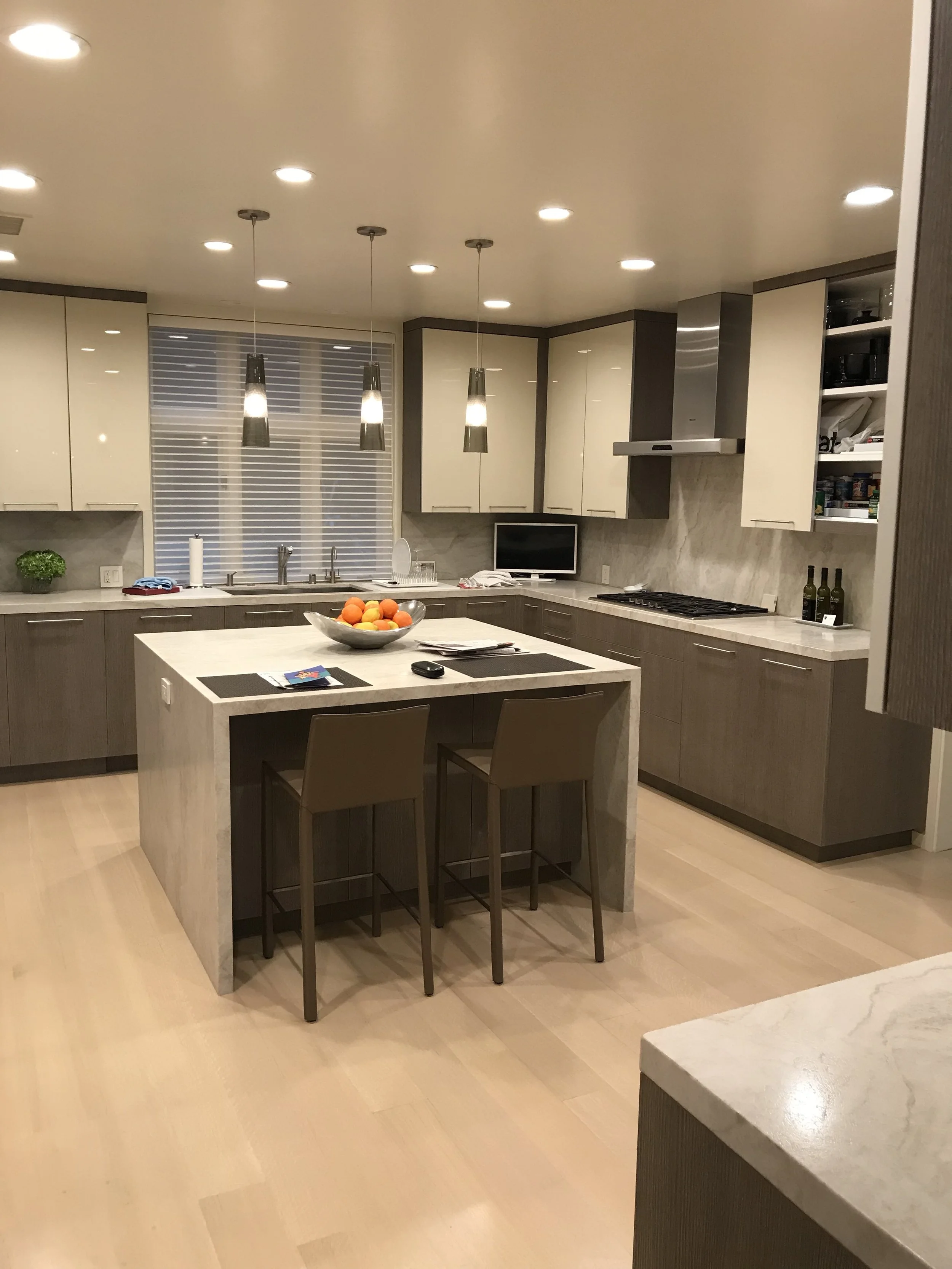 Modern kitchen with beige and gray cabinetry, marble countertops, a central island with chairs, and pendant lighting.
