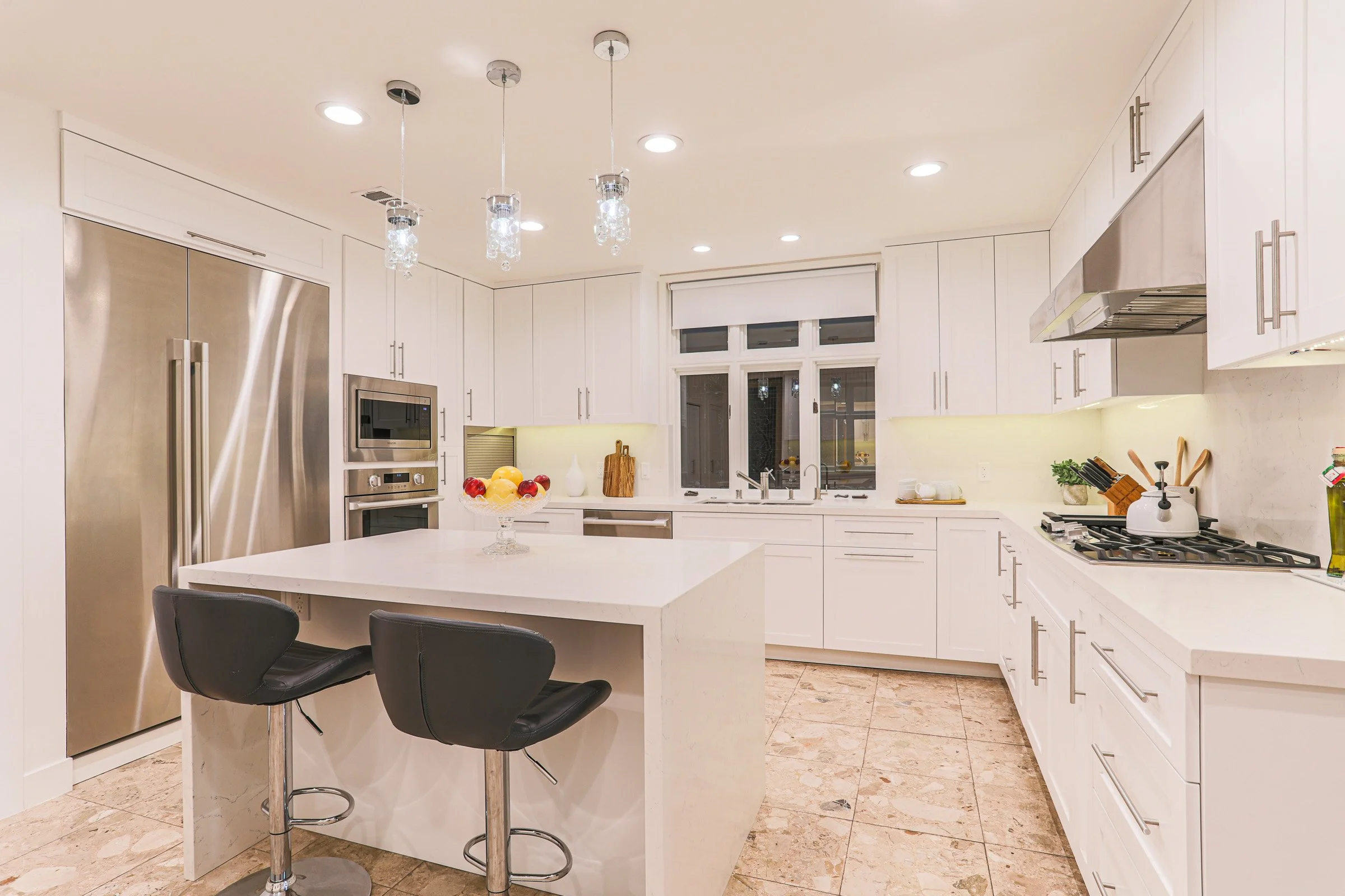 Modern white kitchen with island, black chairs, stainless steel appliances, and beige tile flooring.