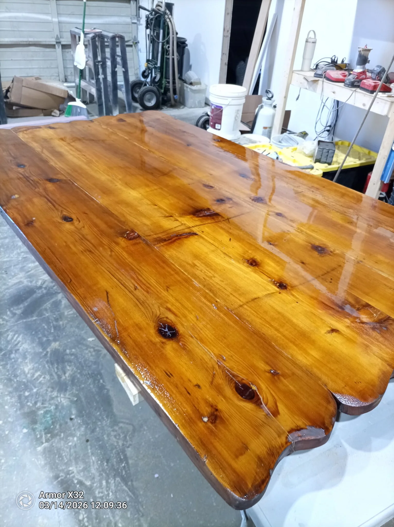 A polished wooden table with a glossy finish in a workshop setting. The background shows tools, equipment, and supplies, including a bucket, vacuum, and power tools.