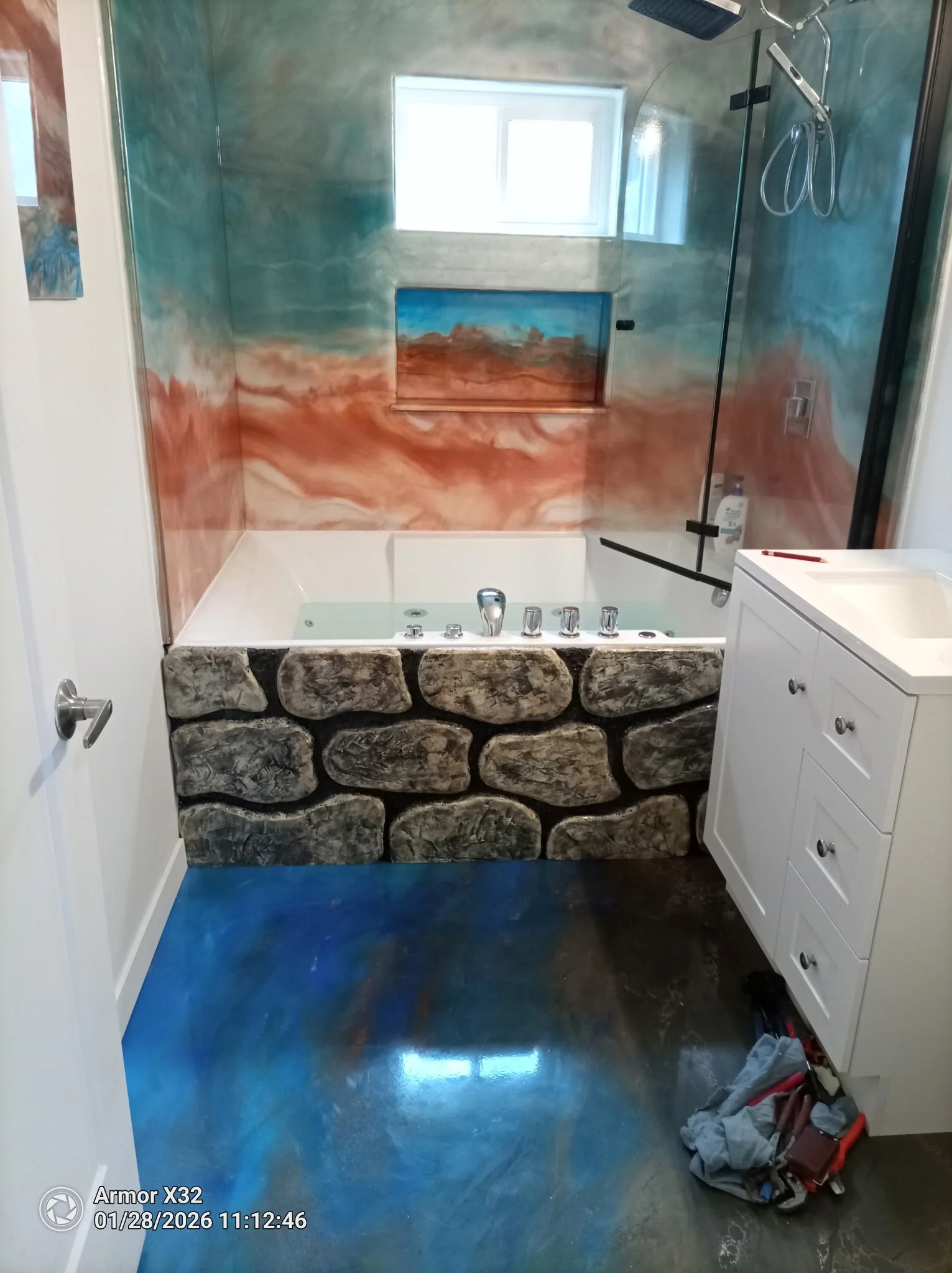 Bathroom with a glass-enclosed shower, painted with a landscape scene of a desert and sky, stone accent on the bathtub, white vanity with drawers on the right, and tools on the floor.