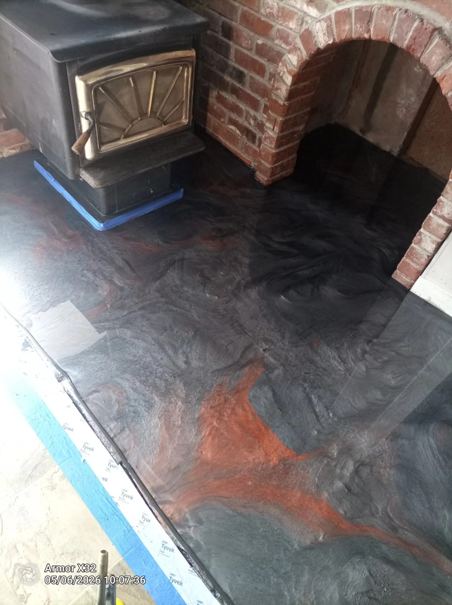 A black stove with a metal door and handle is placed near a brick fireplace with an arched opening. The floor is dark with a reddish-brown streak, and there is insulation material and construction tape visible at the bottom left corner.