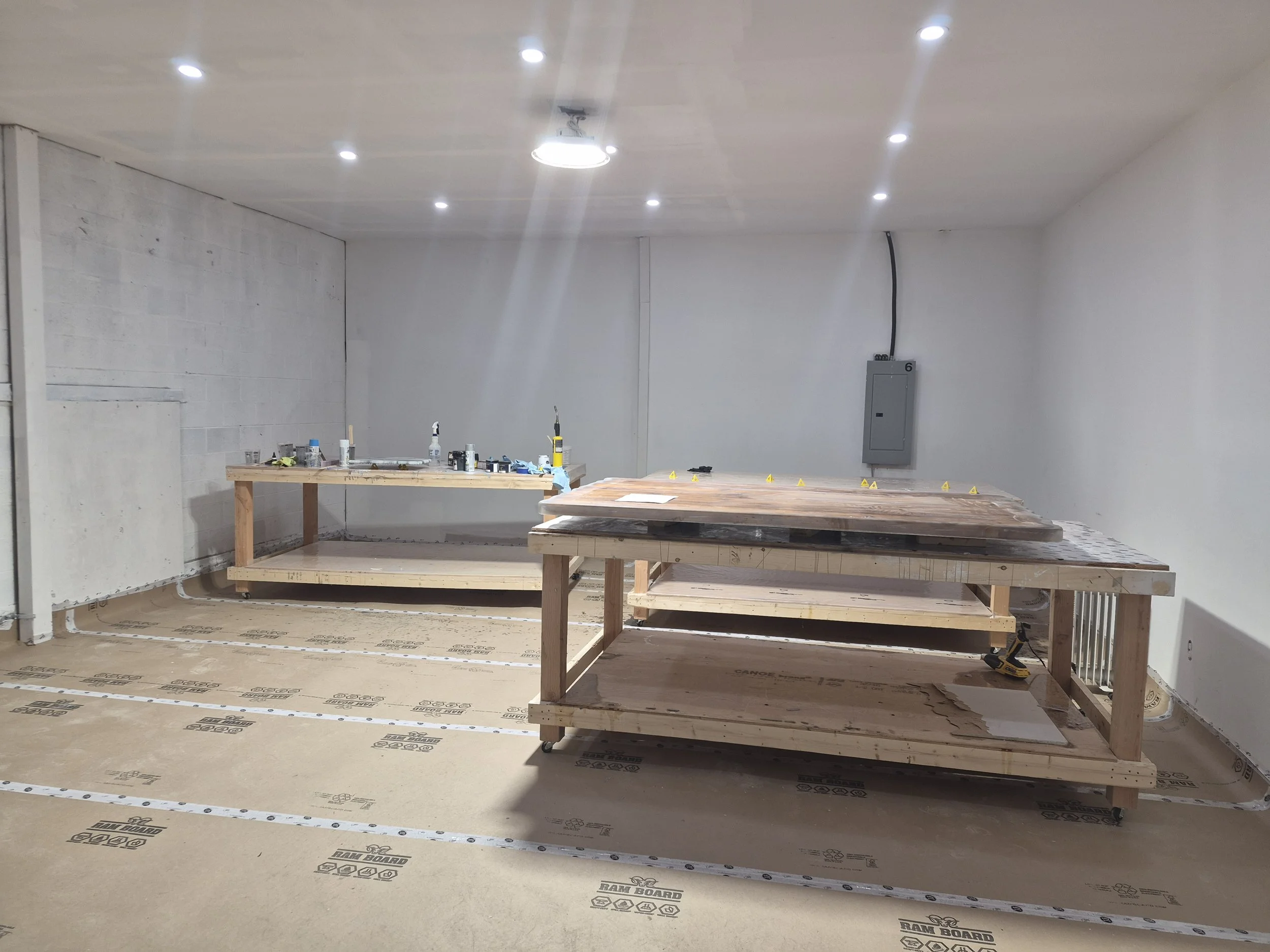 Interior of a room under construction with two wooden workbenches, tools, and construction materials on the surfaces. The walls are white, and the ceiling has multiple recessed lights.